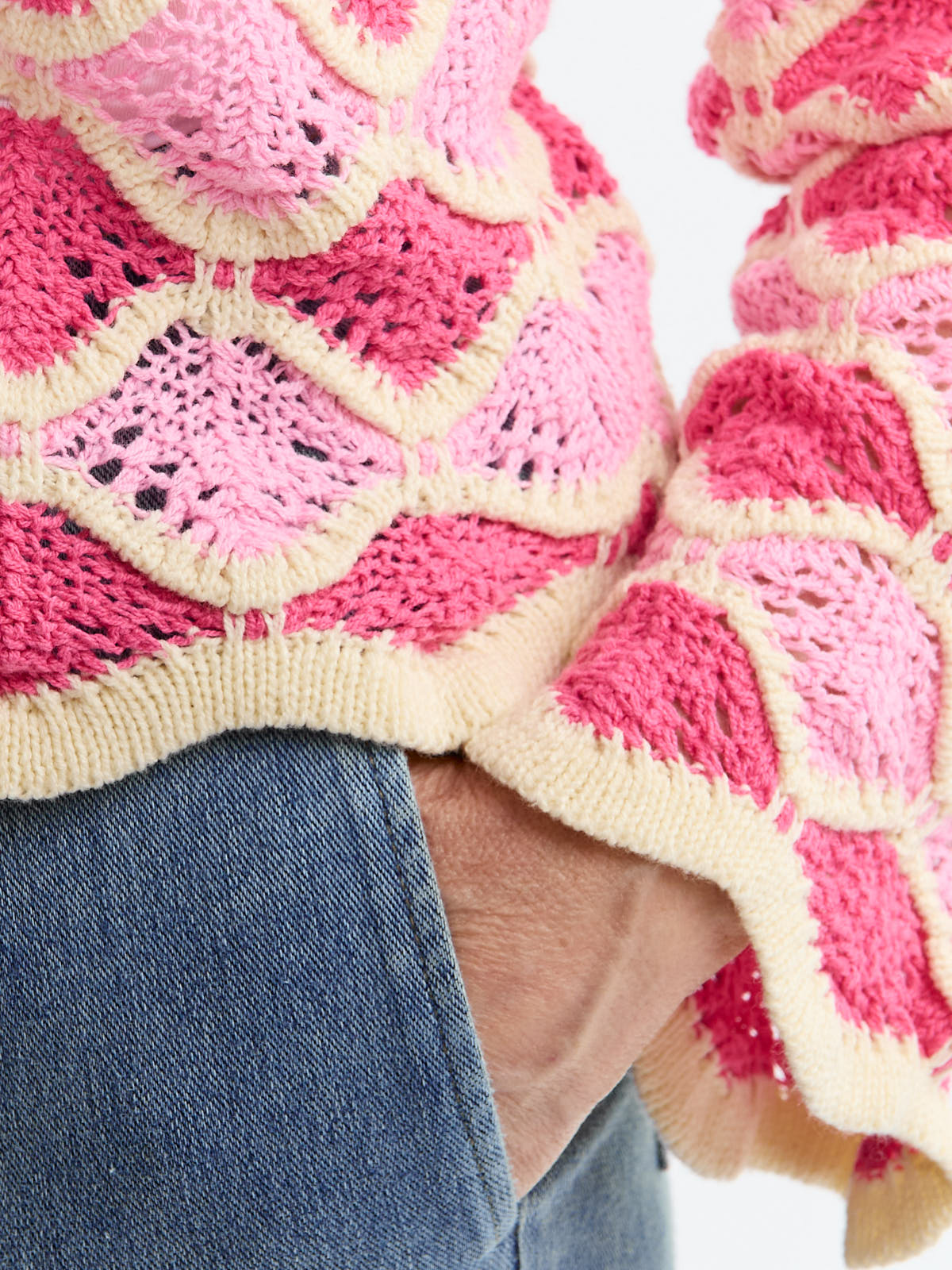 Close-up of a hand in a denim pocket wearing a pink and cream crocheted sweater with diamond patterns.