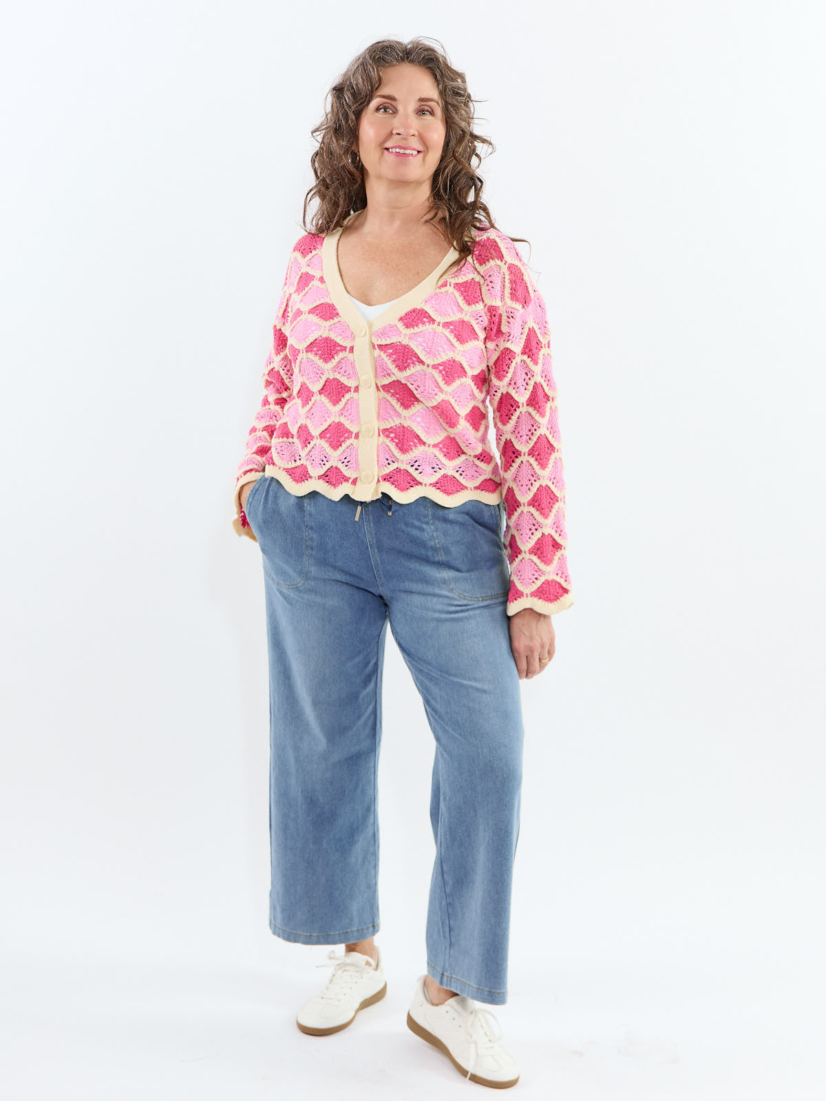 Curly-haired individual in a pink patterned knitted cardigan and blue wide-leg jeans with white sneakers.