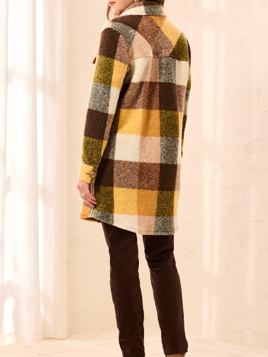 Long plaid coat in yellow, brown, and cream tones paired with dark brown pants in a softly lit room.