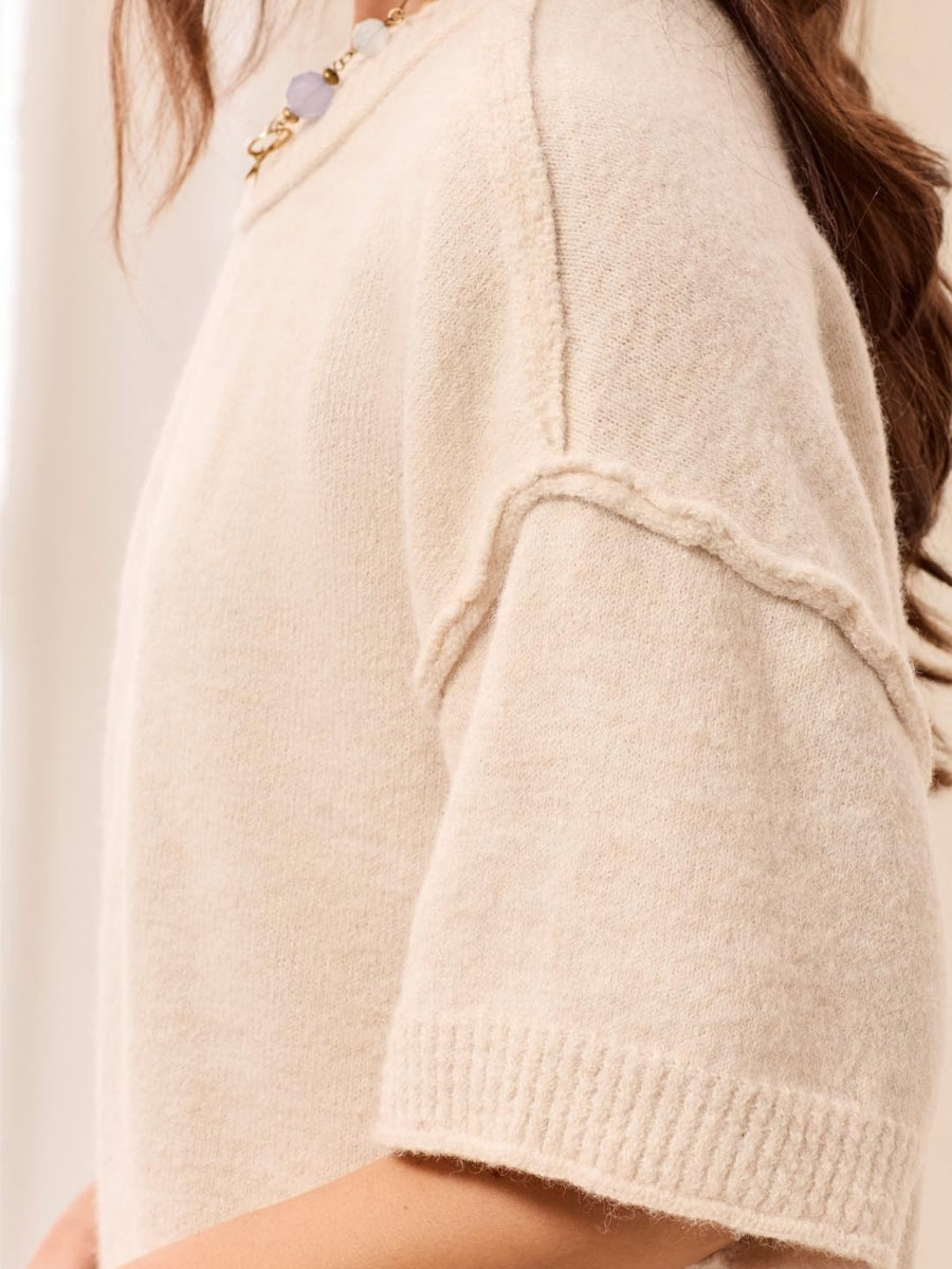Close-up of a cream-colored short sleeve sweater with textured seam detail and ribbed cuffs, paired with a beaded necklace.
