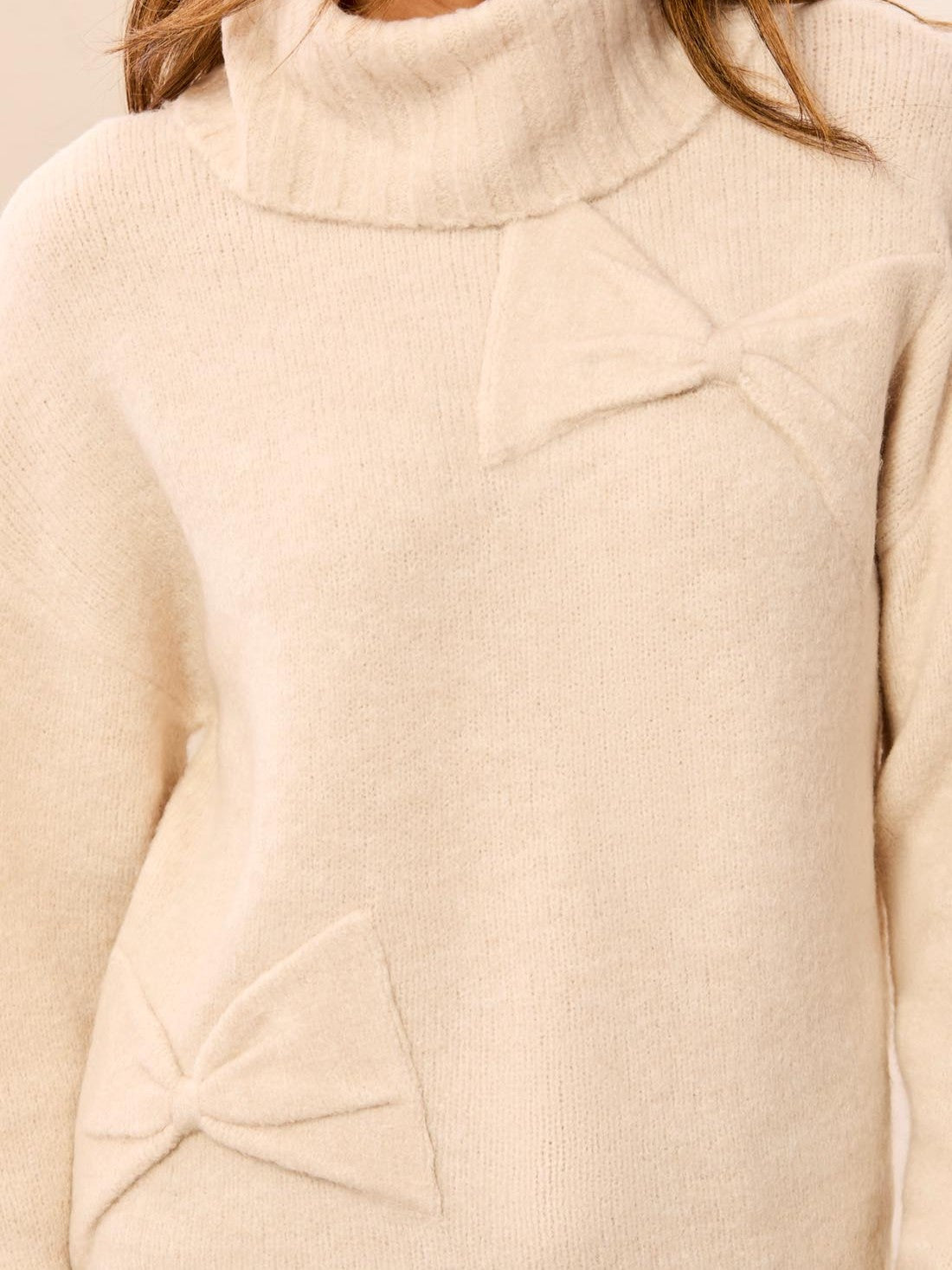 Cream turtleneck sweater with decorative bow details on the chest and lower front