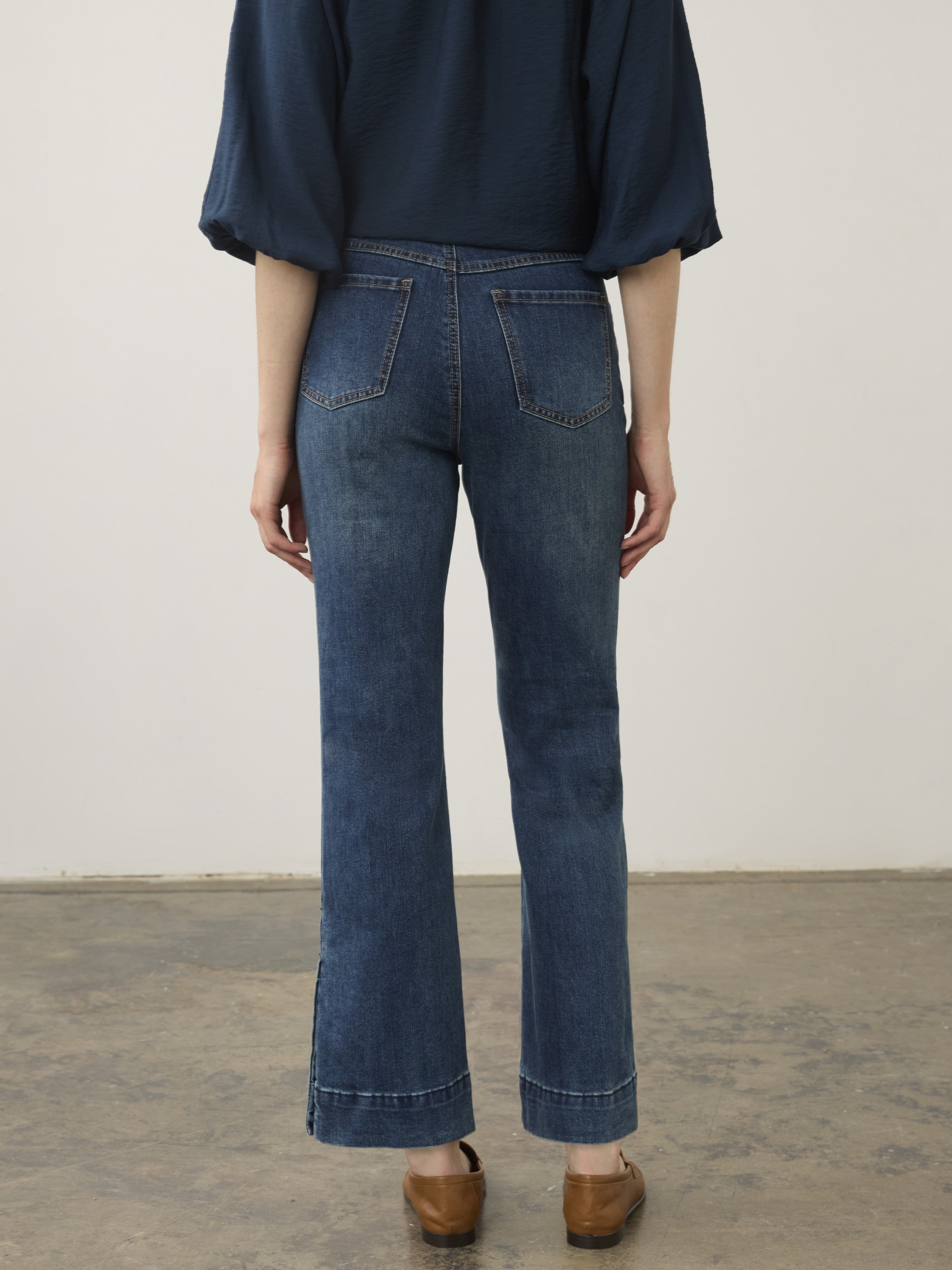 Back view of dark blue denim jeans with a slight flare and side slits paired with brown shoes.