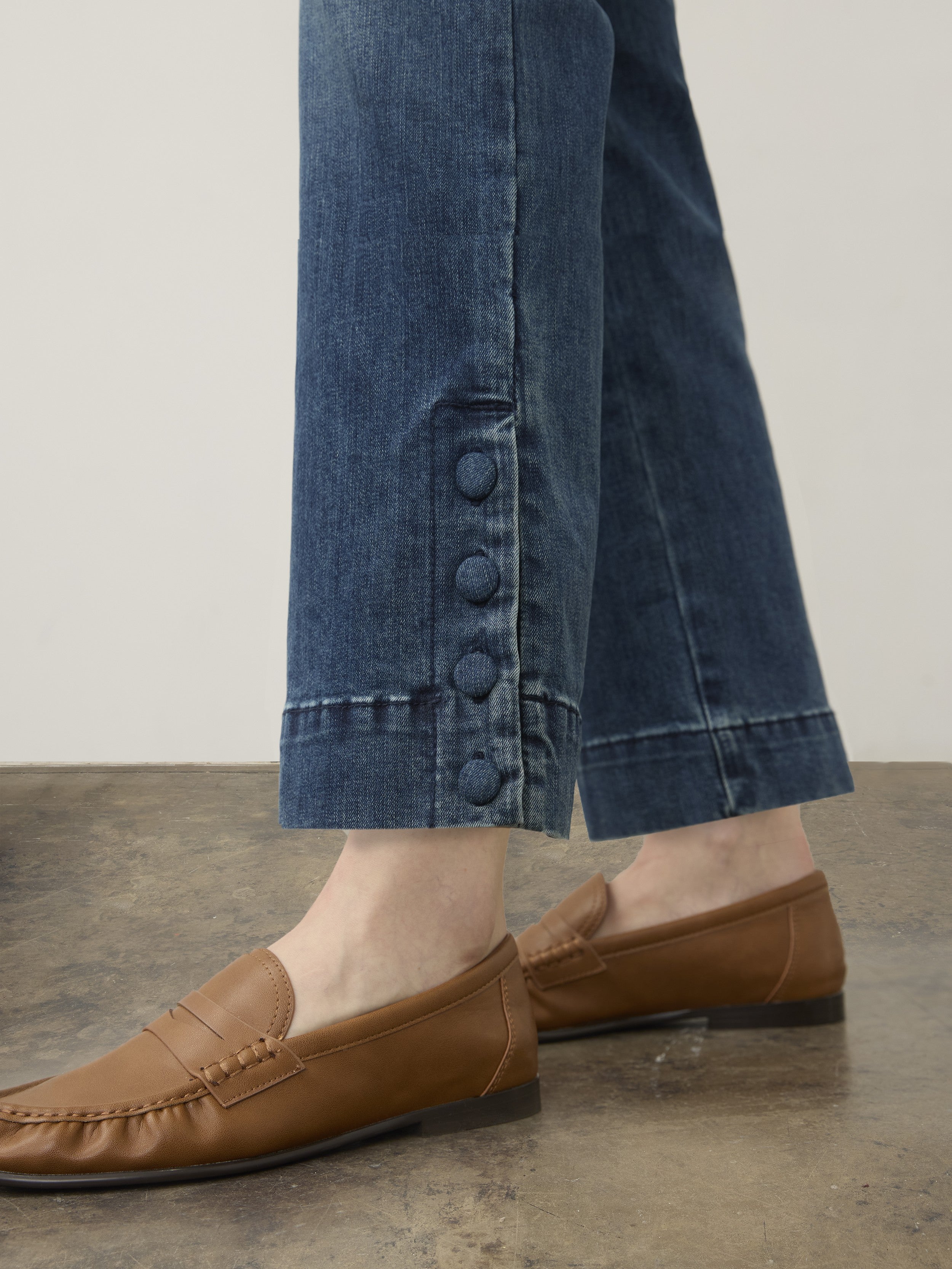 Close-up of denim jeans with four button details near the hem paired with brown leather loafers on a concrete floor.