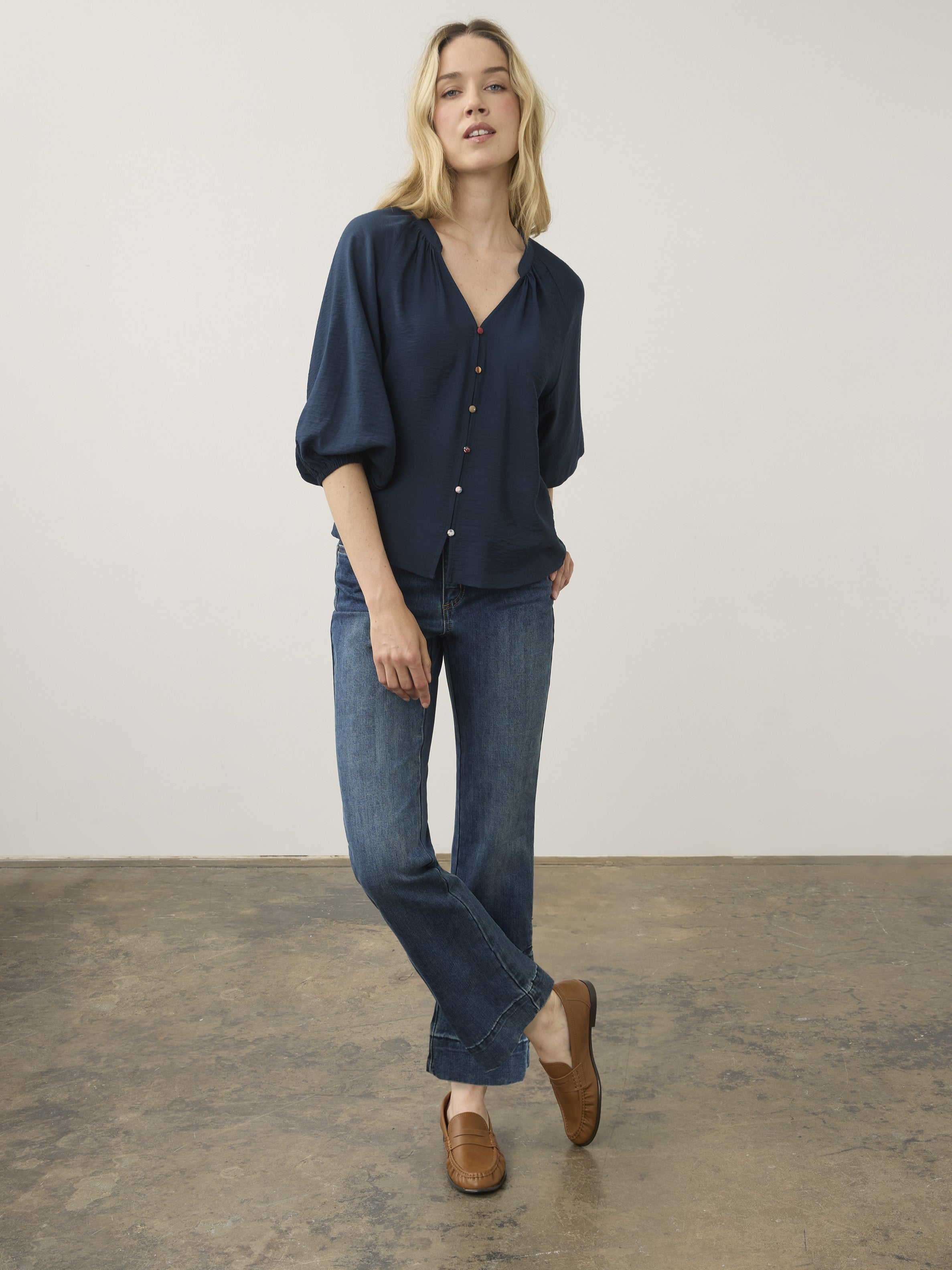 Individual standing on a concrete floor wearing a dark blue buttoned blouse, blue denim jeans, and tan leather loafers.