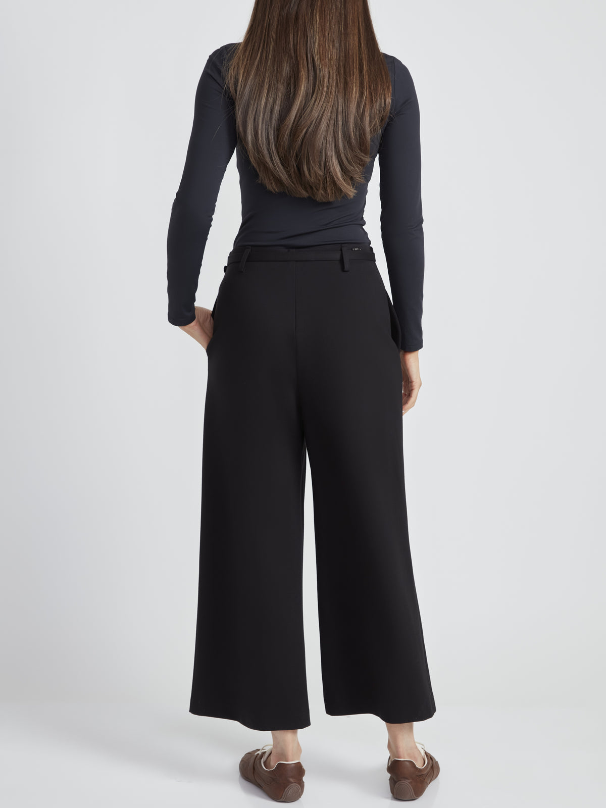 Back view of a figure in a long-sleeve black top and wide-leg black pants against a plain light background.