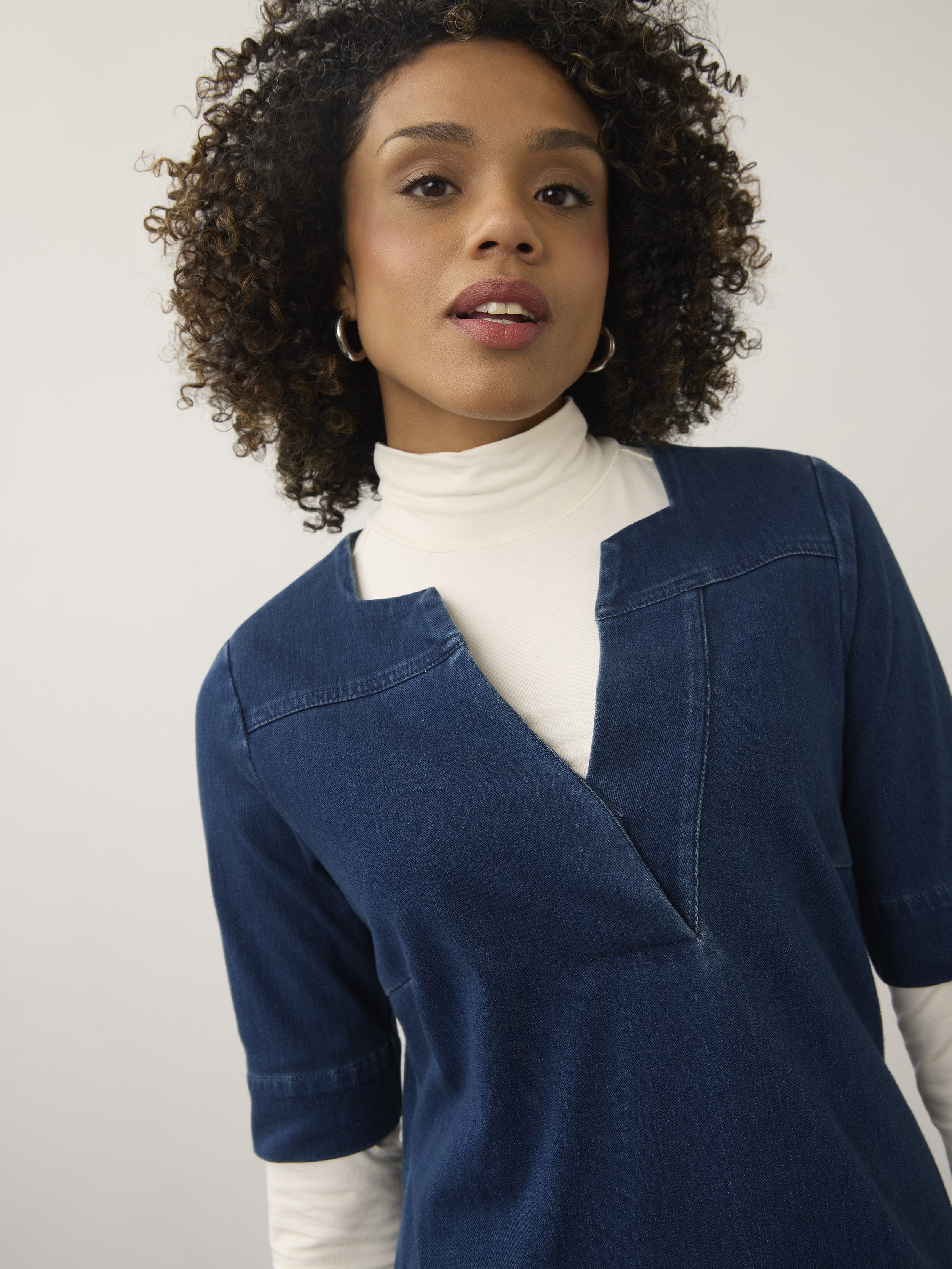 Close-up of a model wearing a dark blue denim top layered over a white long-sleeve turtleneck against a plain background.
