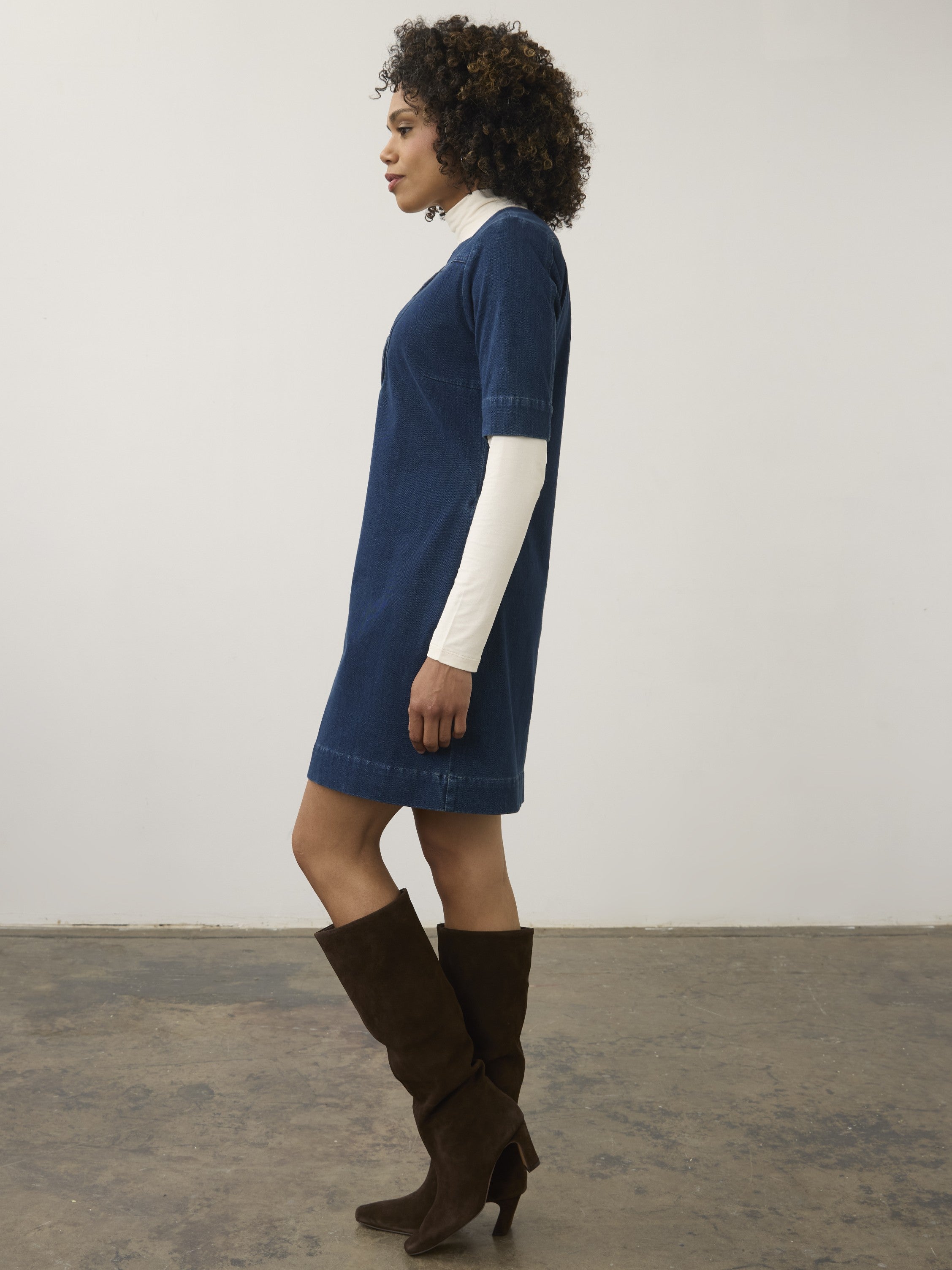 Side view of a short-sleeve denim dress over a long-sleeve white top paired with knee-high brown suede boots.