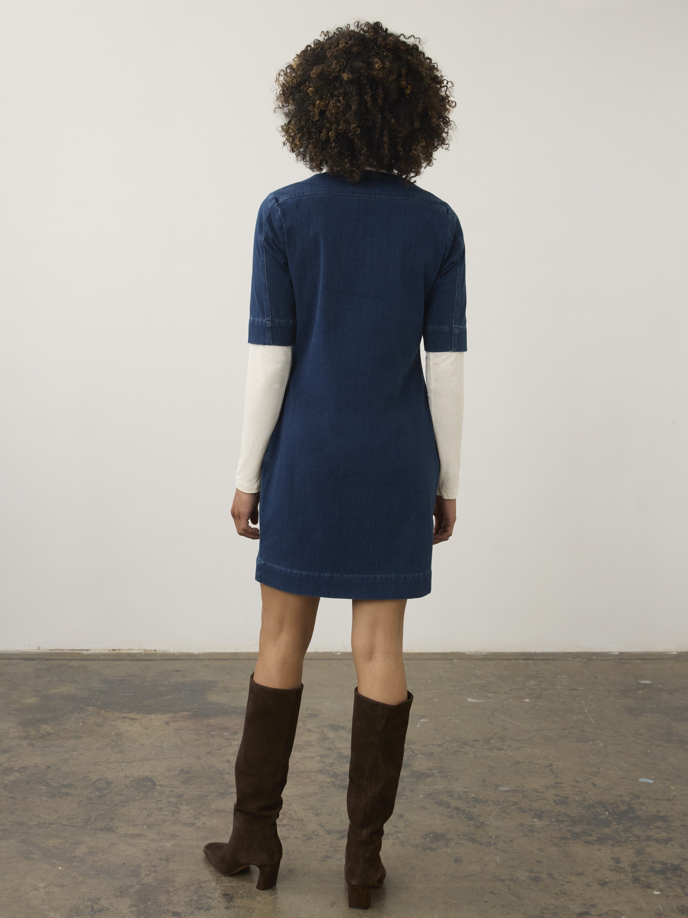 Back view of a knee-length dark blue denim dress with short sleeves over a white long sleeve top, paired with dark brown boots.
