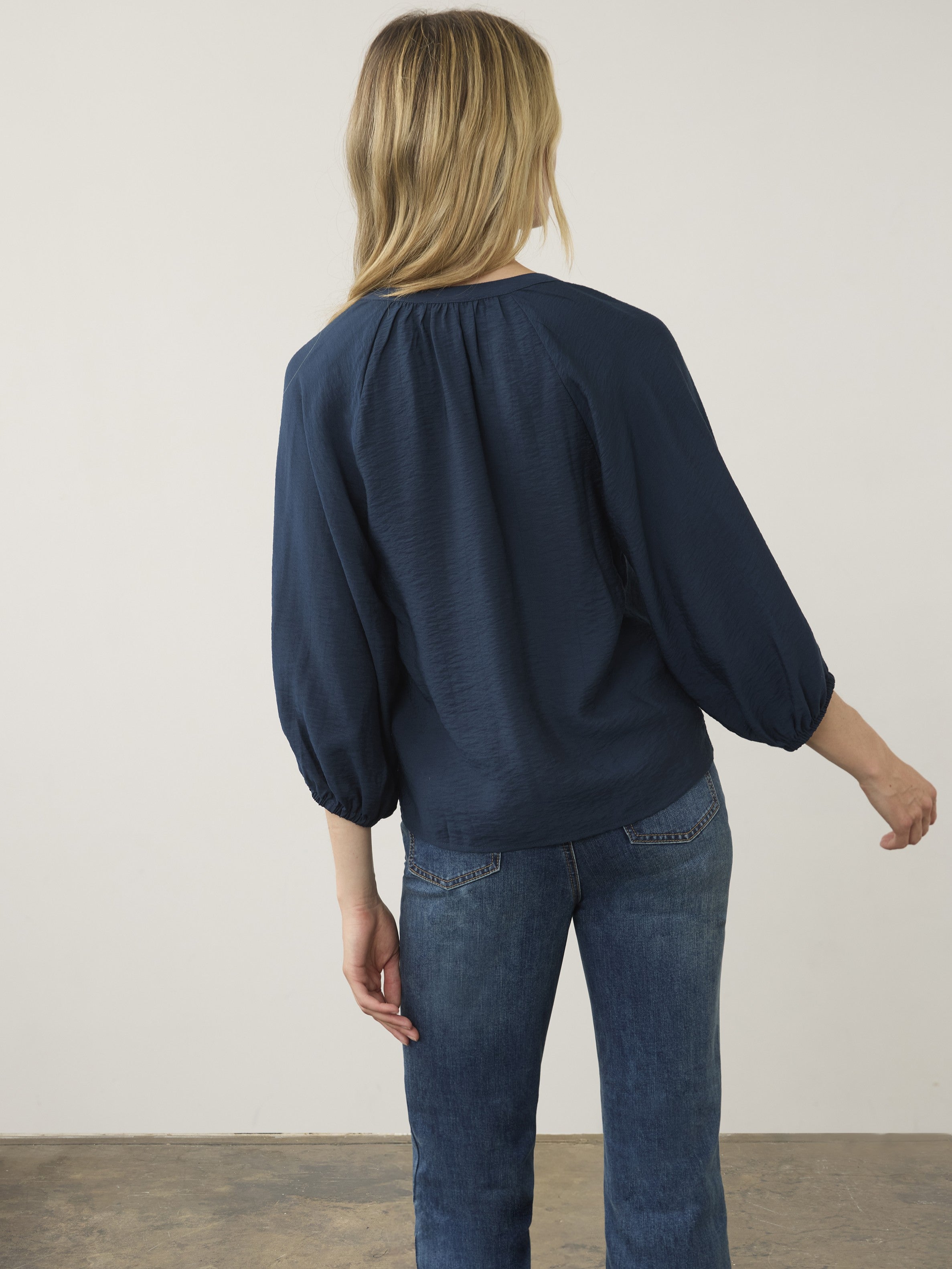Back view of a navy blue long-sleeve blouse paired with medium wash denim jeans against a plain background.