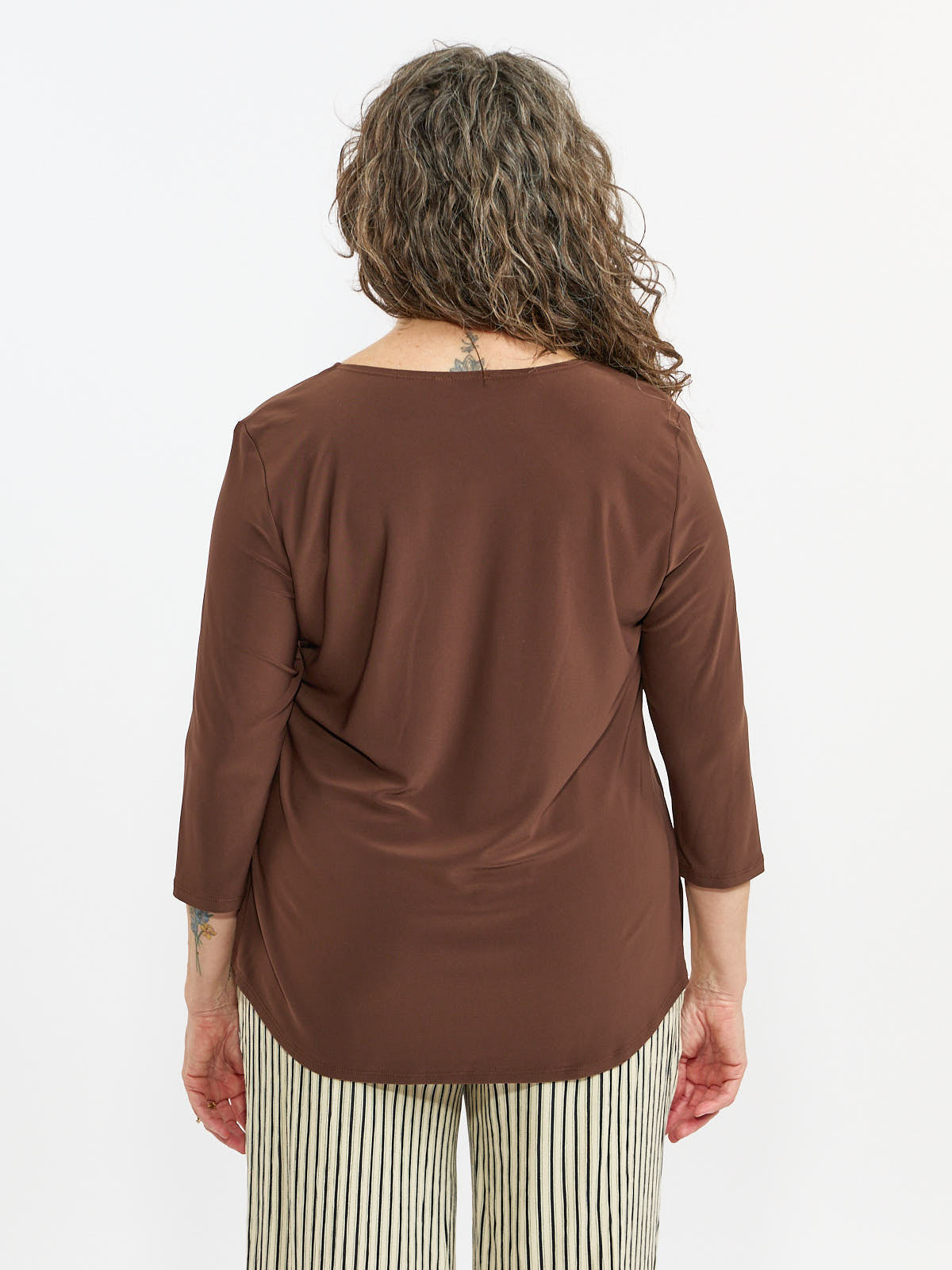 Back view of a person with curly hair wearing a brown long-sleeve top and striped pants against a white background.