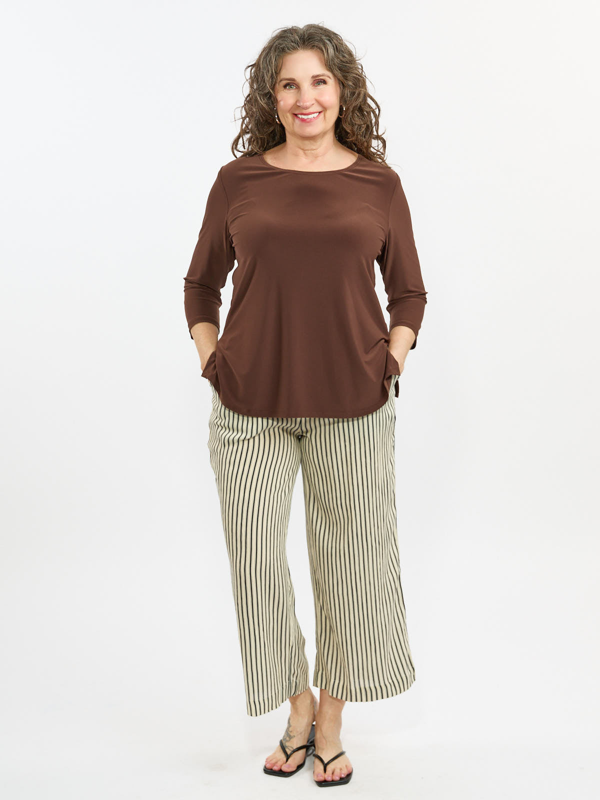 Curly hair with brown long-sleeve top and beige striped wide-leg pants paired with black sandals on white background.