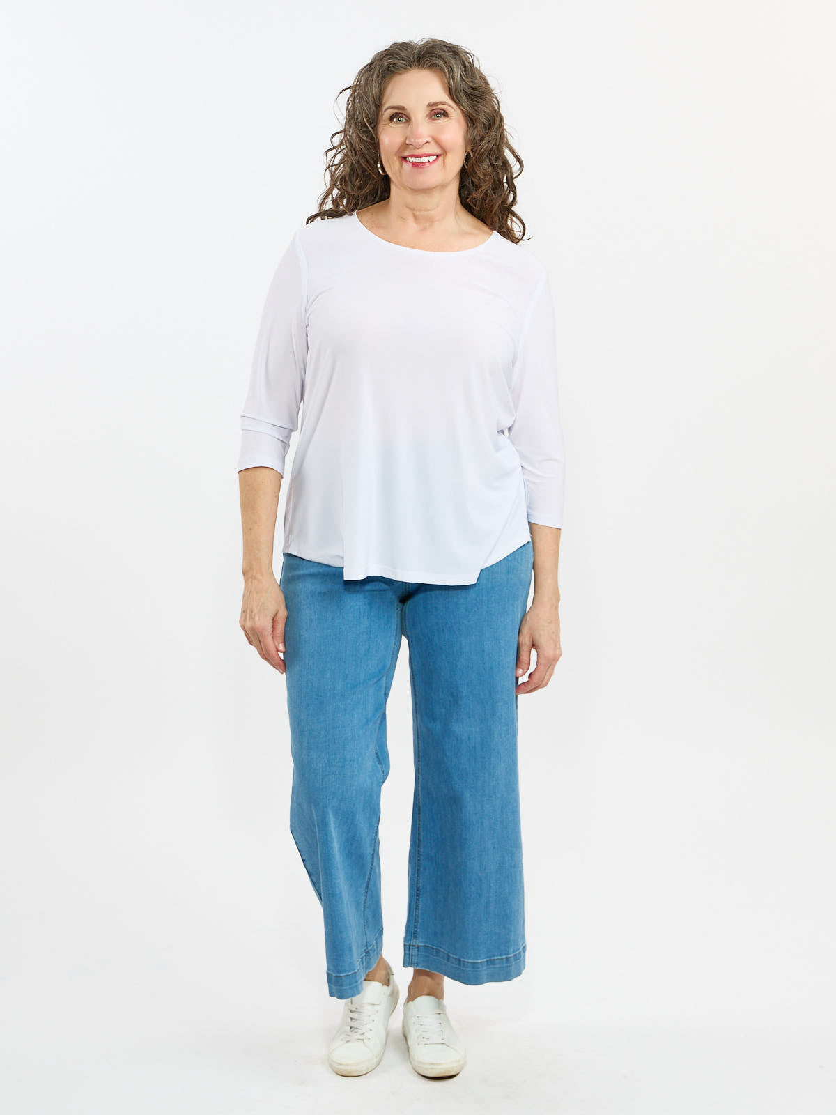 Person standing in a white long-sleeve top, wide-leg blue denim jeans, and white sneakers against a white background