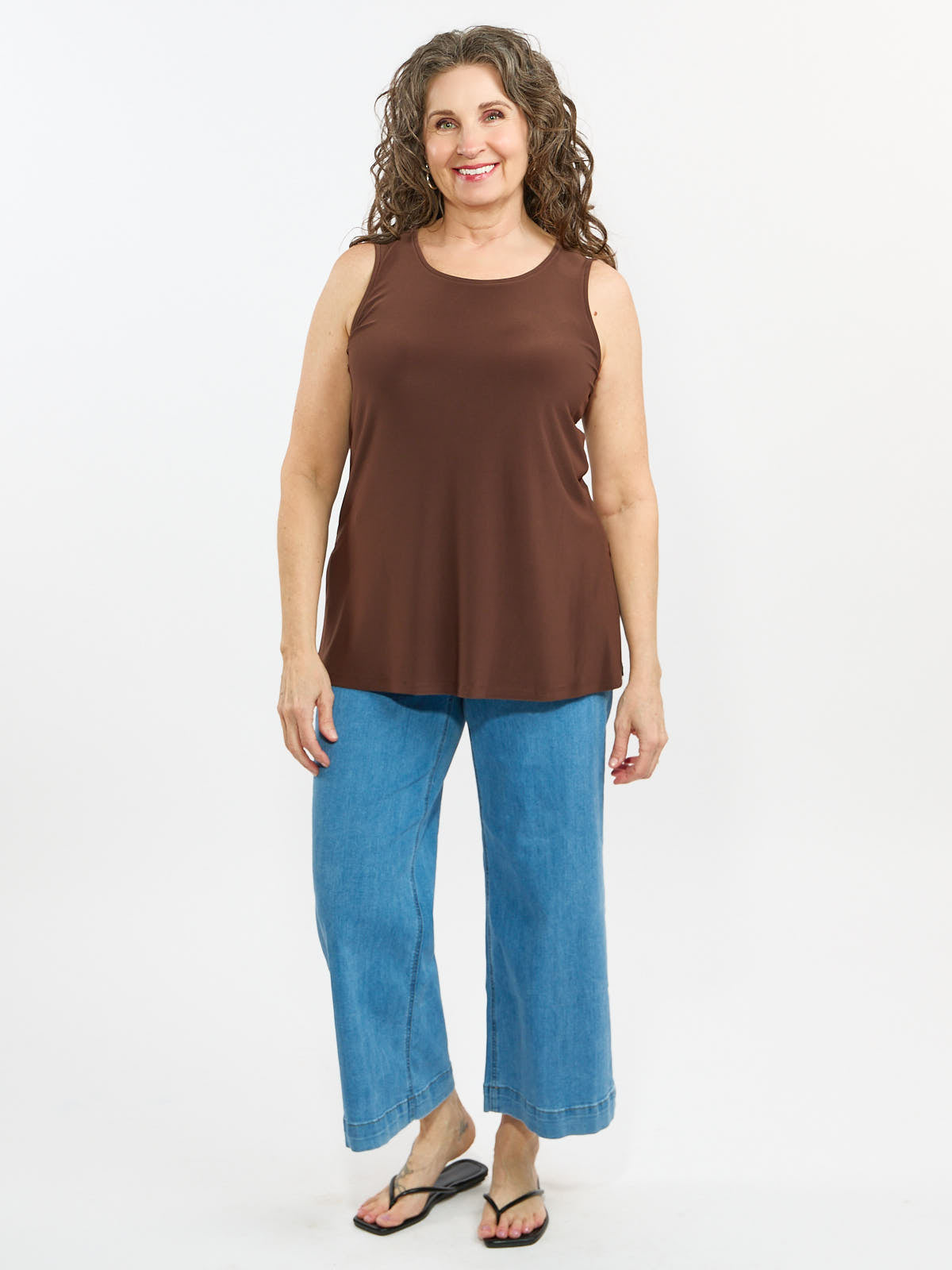 Smiling individual wearing a sleeveless brown top and wide-leg blue denim pants with black sandals on white background.