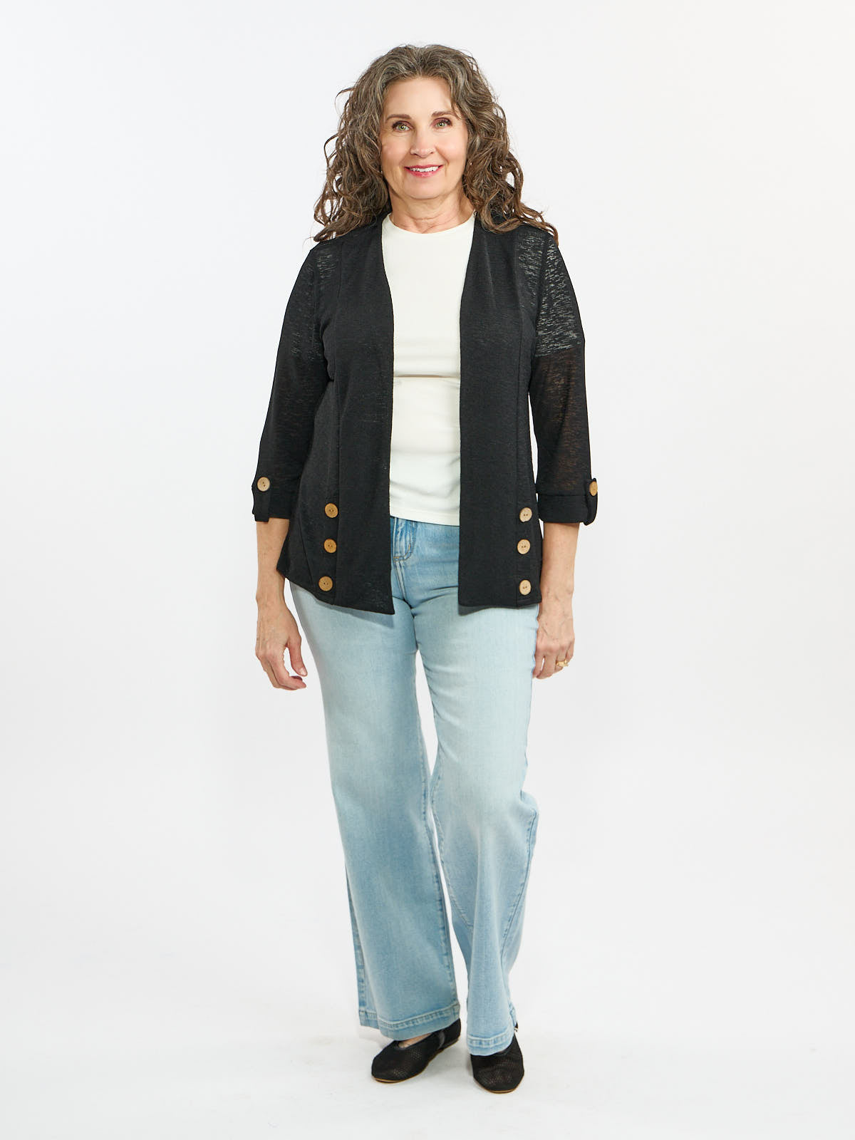 Curly gray hair individual in a black buttoned cardigan over white top and light blue jeans on white background