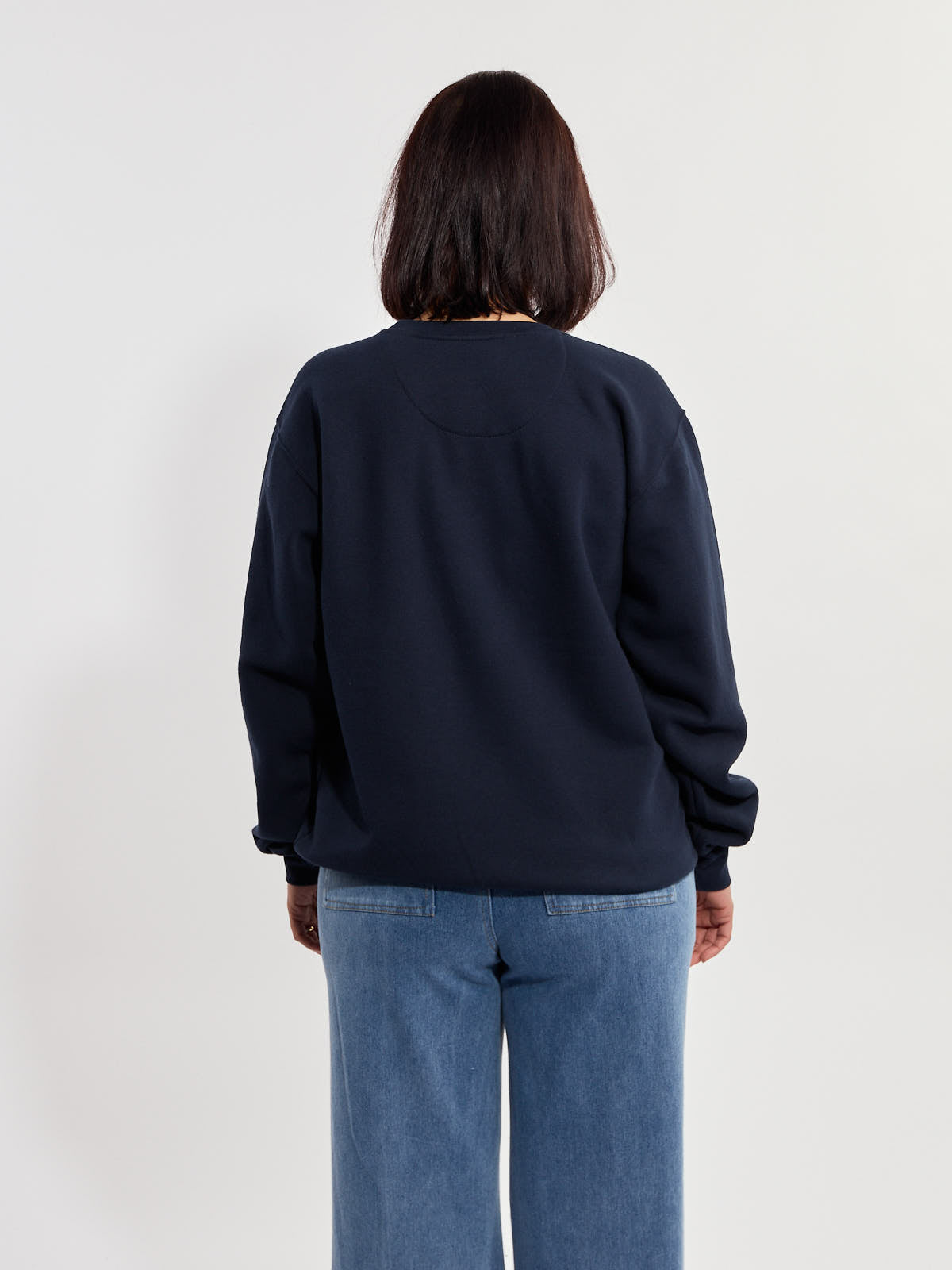 Back view of a person wearing a navy blue sweatshirt and light blue denim jeans against a plain background.