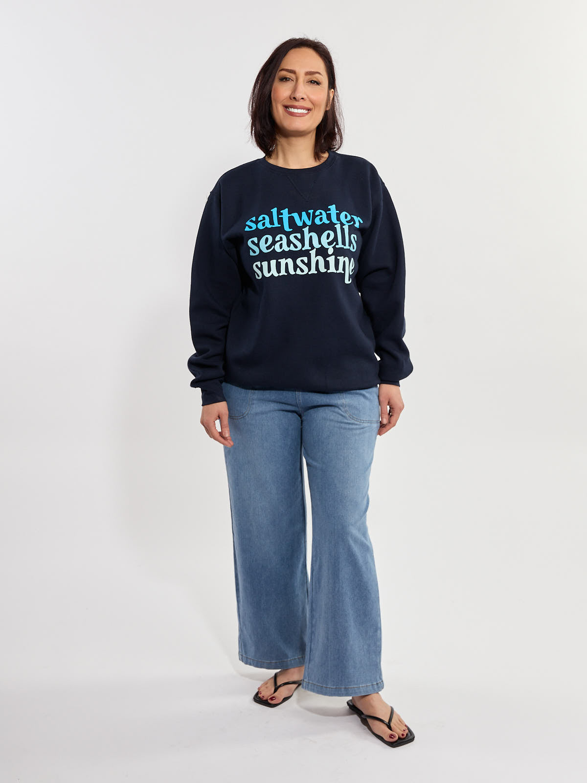 Navy blue sweatshirt with "saltwater seashells sunshine" text paired with light blue wide-leg jeans and black sandals.