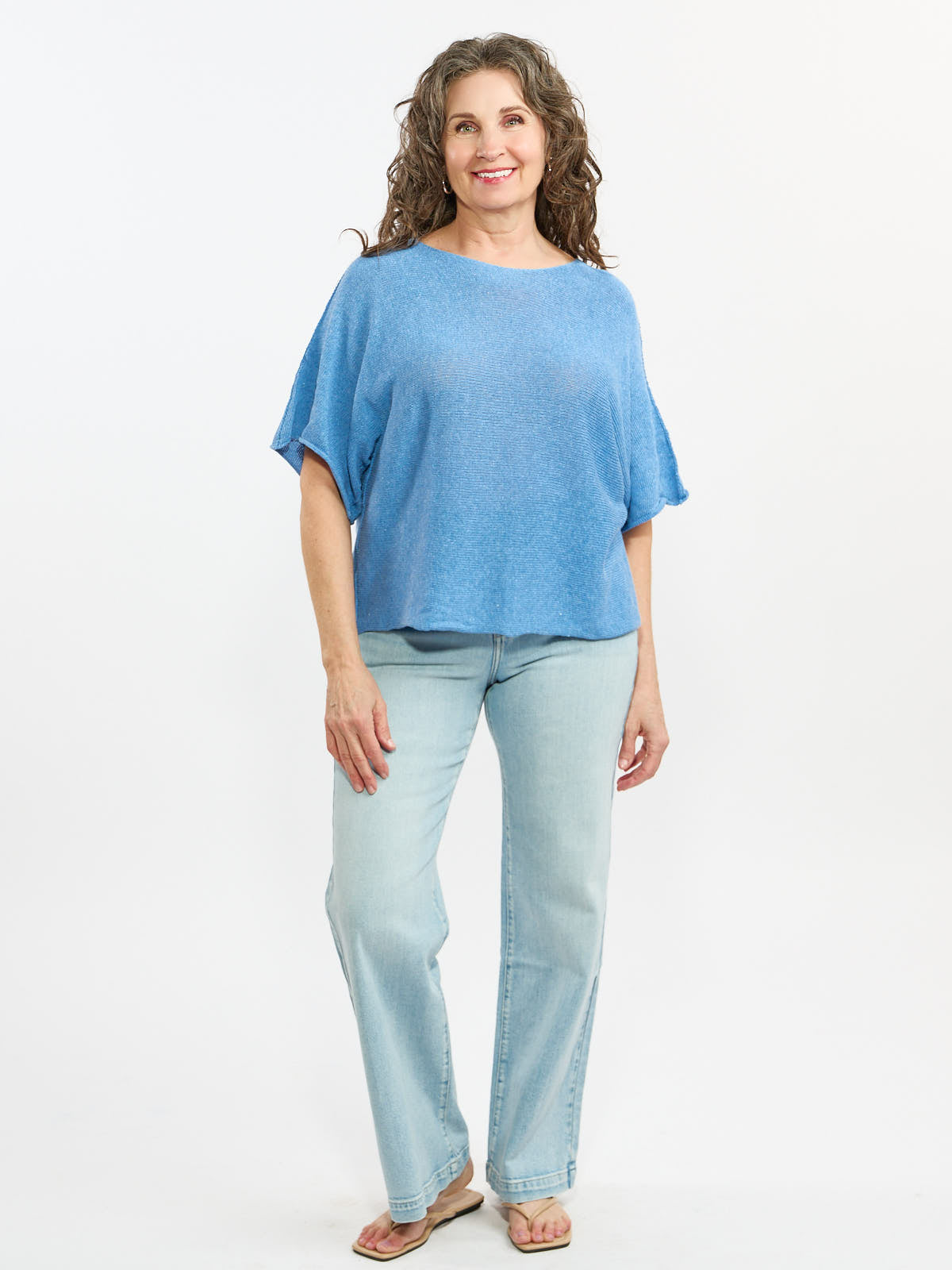 Person standing wearing a loose blue short-sleeve top and light-wash denim jeans against a white background.