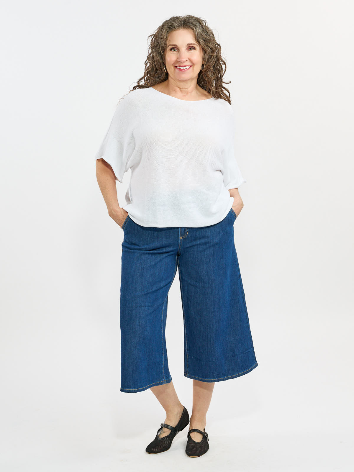 Smiling figure in a loose white top and wide-leg blue denim cropped pants with black flat shoes on a white background.