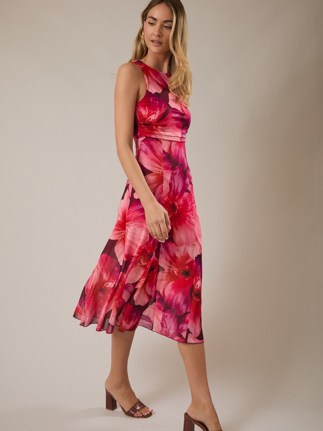 Model in a sleeveless, knee-length dress with large pink and red floral print and brown open-toe heels