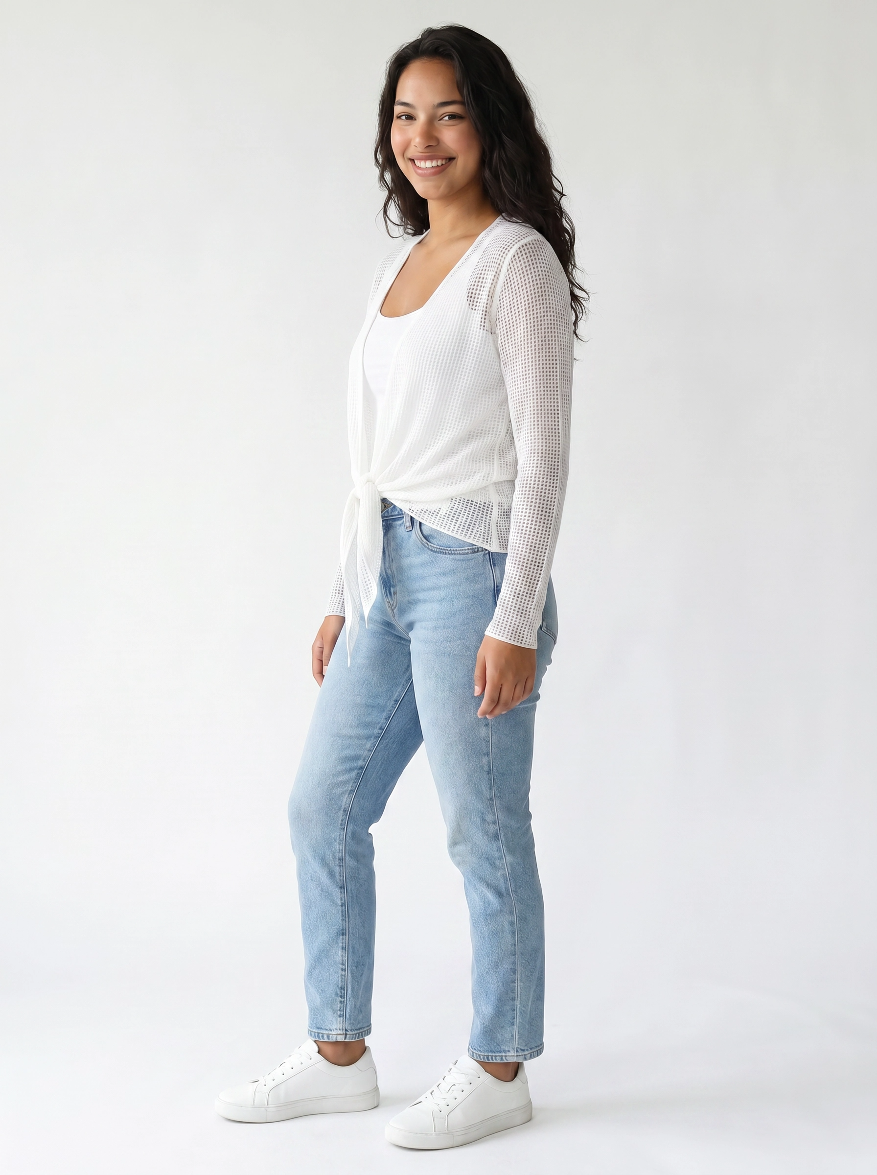 Young individual in light blue denim jeans, white tank top, and white knit long-sleeve top tied at the waist with white sneakers.