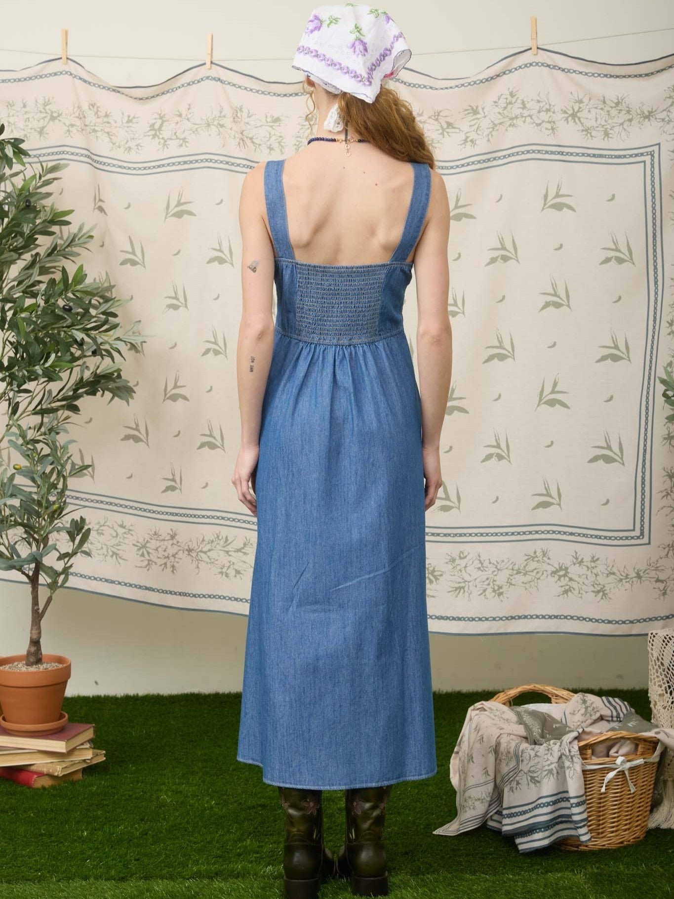 Back view of a blue denim sleeveless dress with a smocked bodice and wide straps against a floral backdrop.