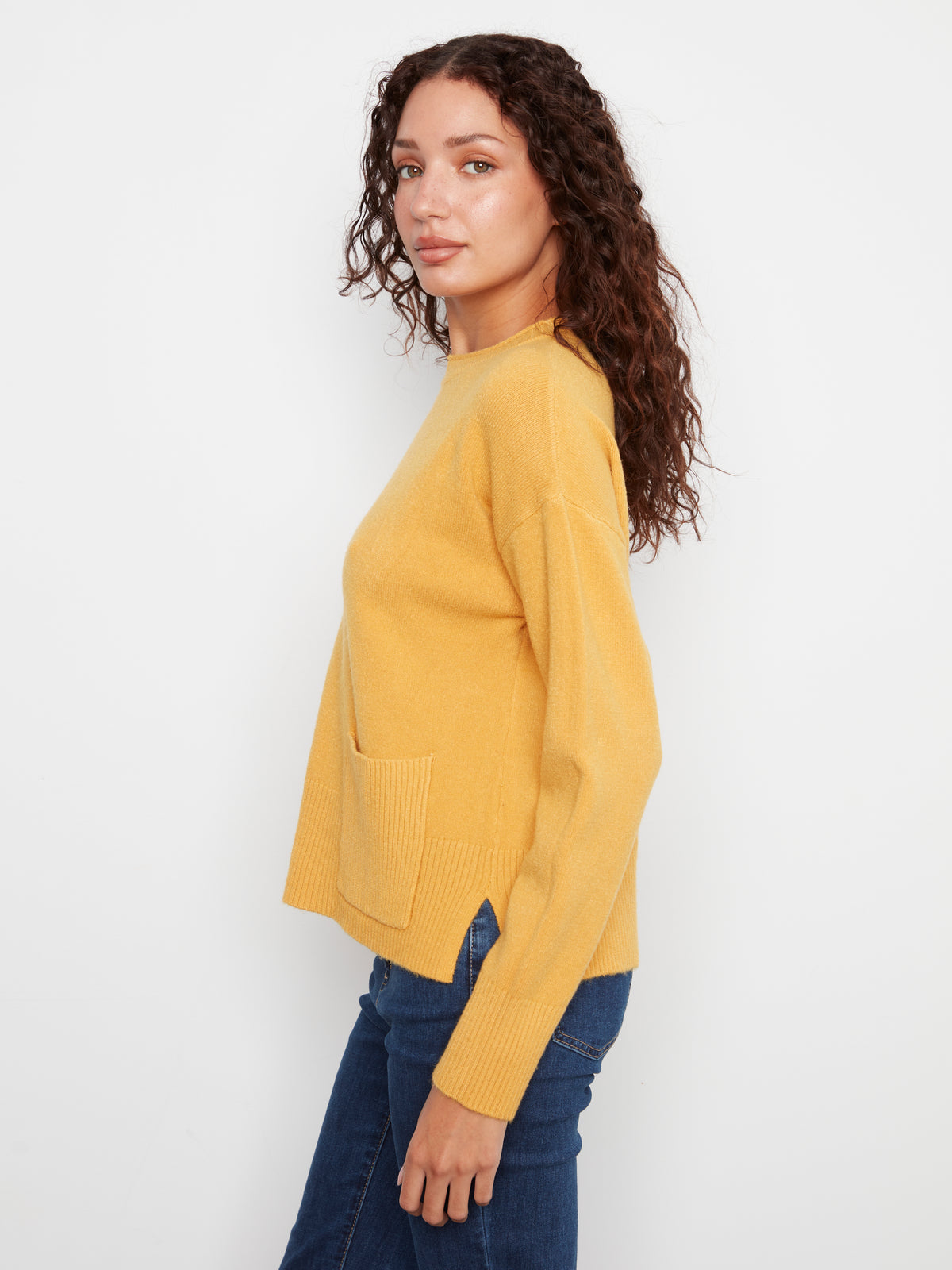 Curly-haired model in a mustard yellow sweater with a front pocket and dark blue jeans