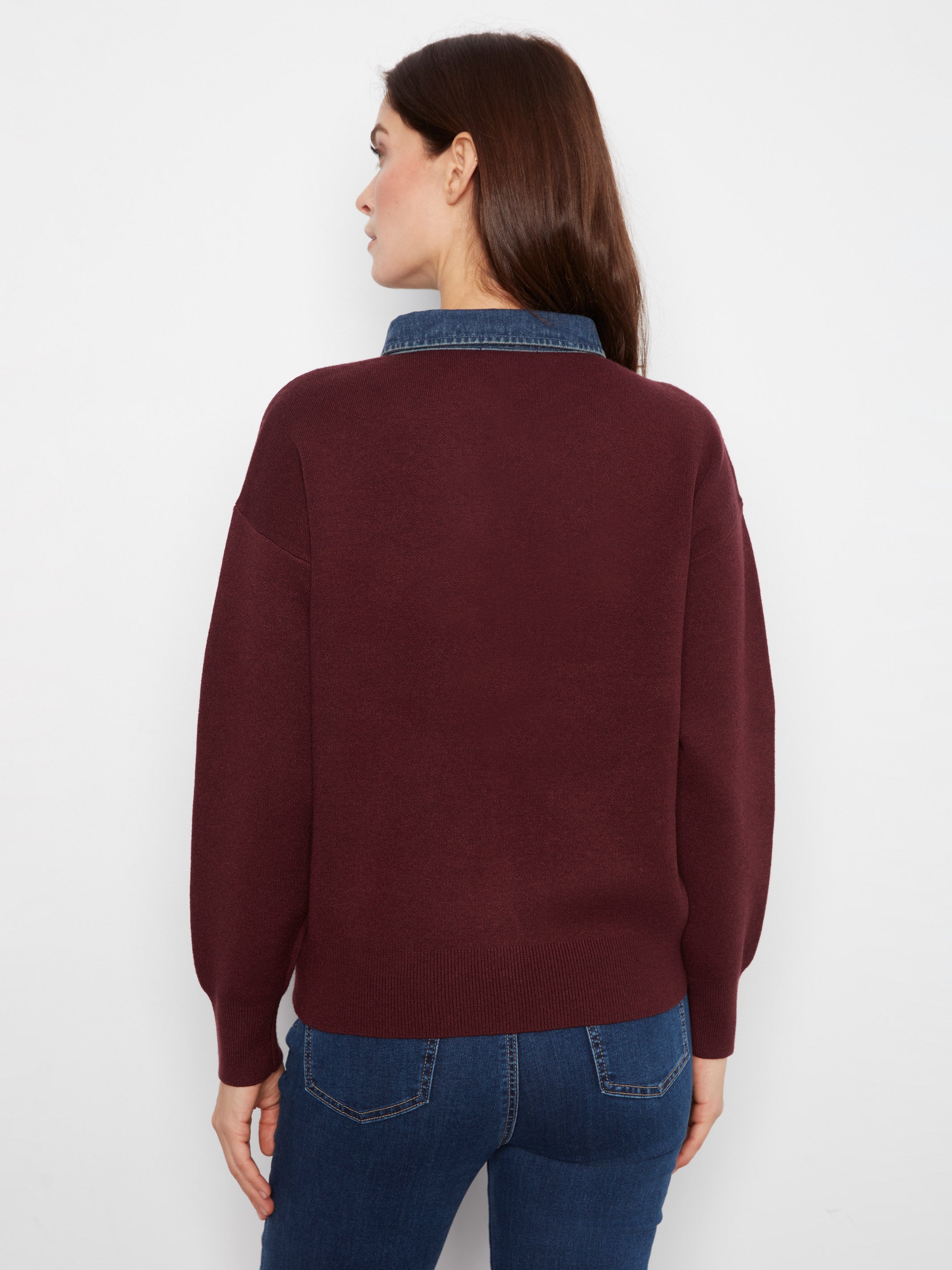 Back view of a dark burgundy sweater layered over a blue denim shirt with blue jeans against a white background.