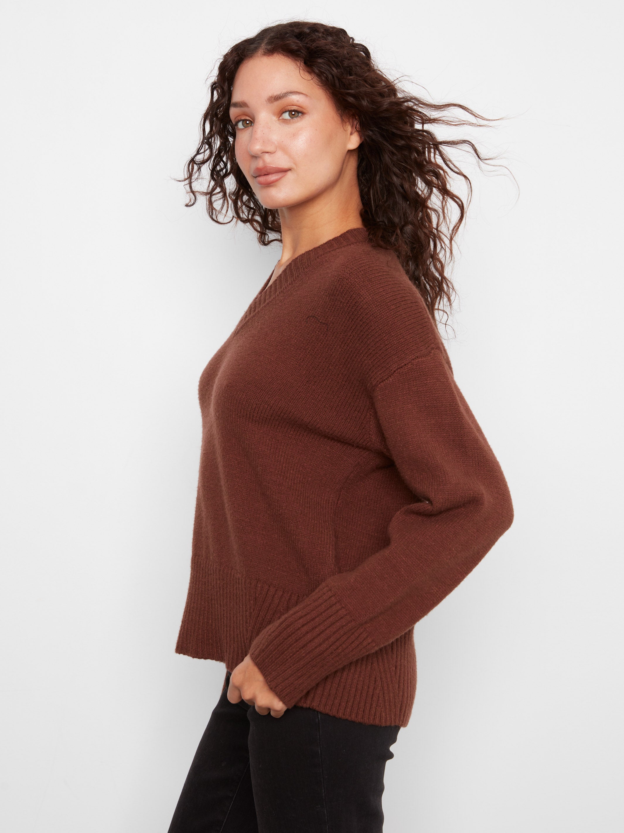 Side view of a brown knitted sweater with ribbed cuffs and hem paired with black pants against a white background.