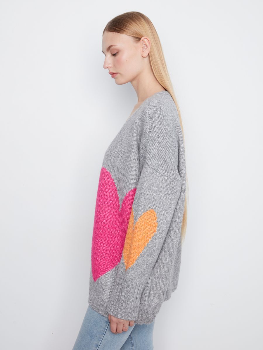 Side view of a grey knitted sweater with large pink and orange heart patterns worn with light blue denim jeans.