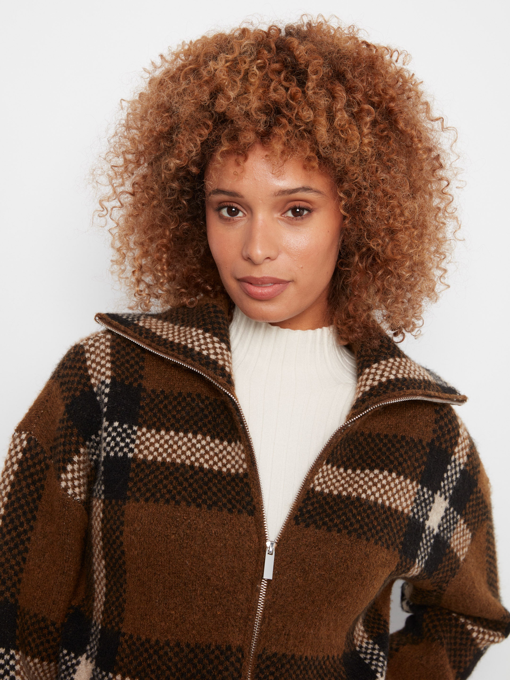 Close-up of a curly-haired figure in a brown and black plaid zip-up jacket over a white ribbed top.