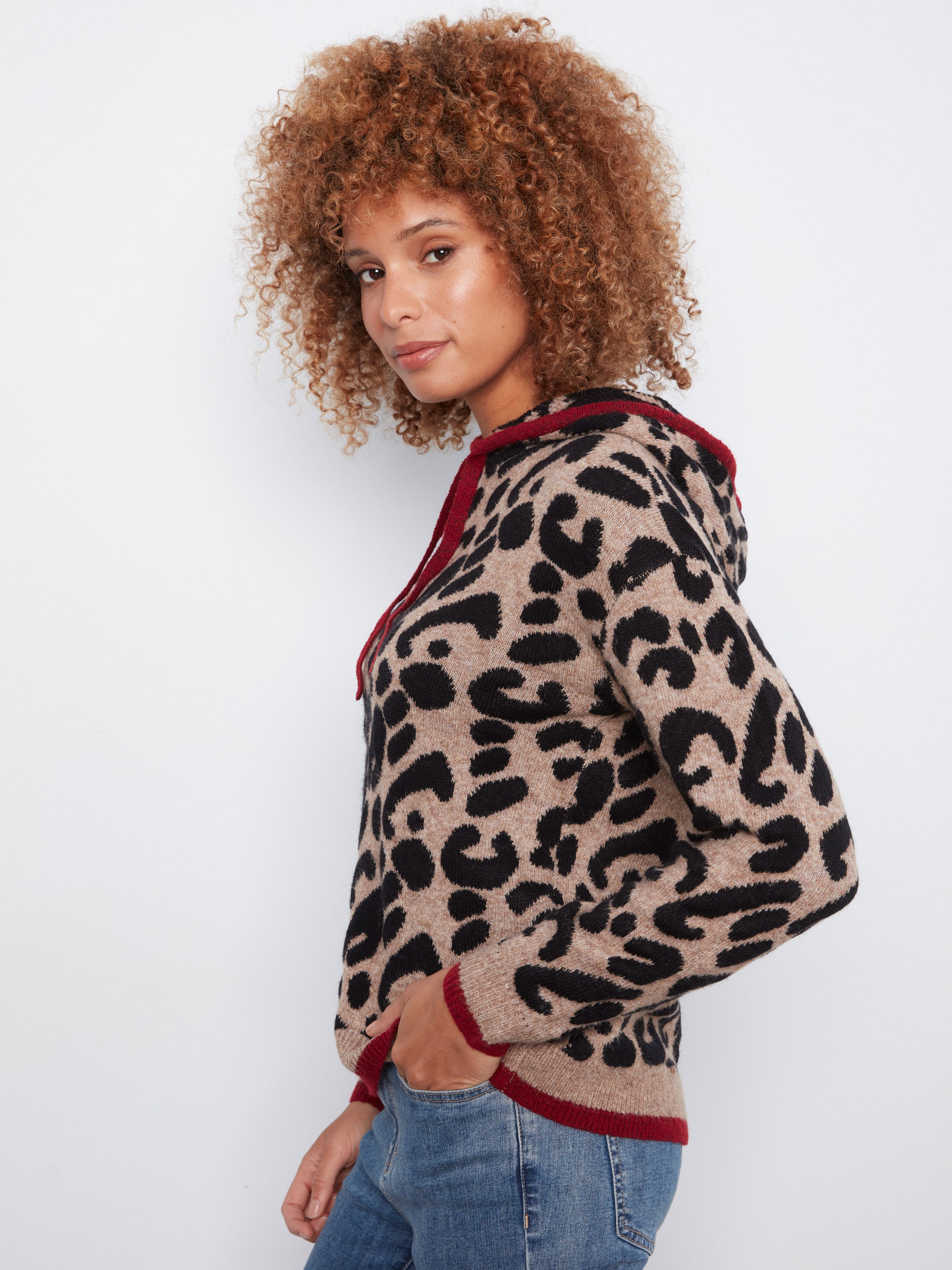 Curly hair model in a tan and black leopard print sweater with red trim and blue denim jeans against white background.