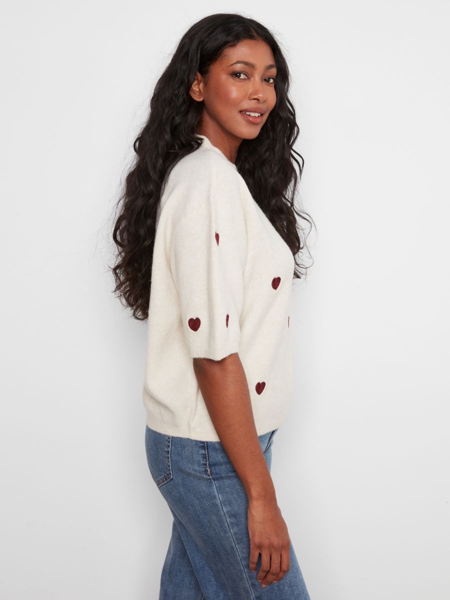 Side view of a person with long curly hair wearing a cream sweater with small red heart patterns and blue jeans.
