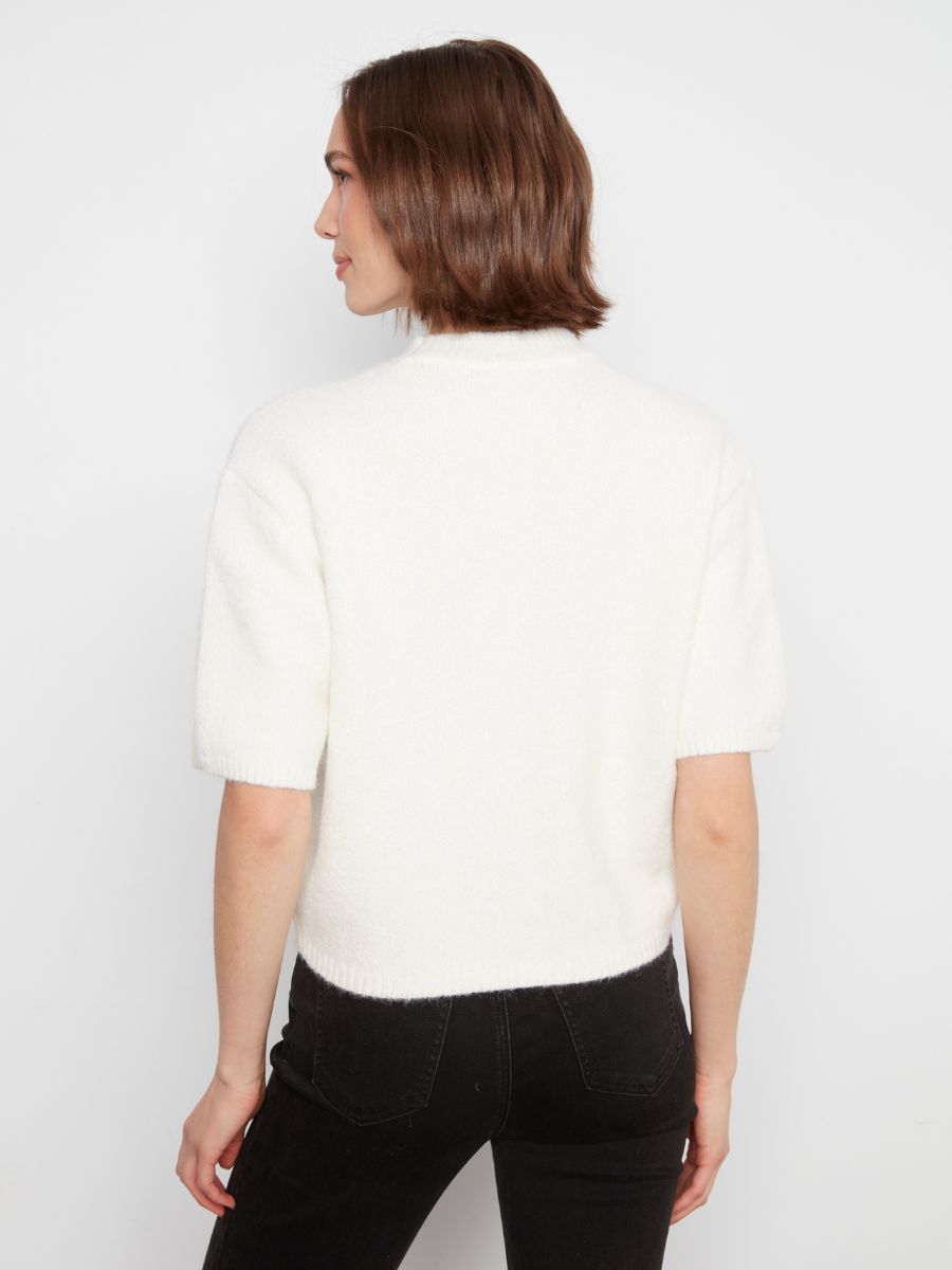 Back view of a cream-colored short-sleeve sweater paired with black denim pants against a white background.