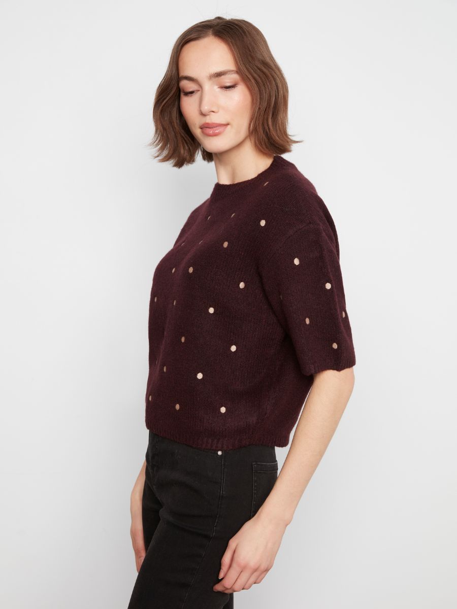 Side view of a dark plum knitted sweater with short sleeves and small contrasting dot patterns worn with black pants.