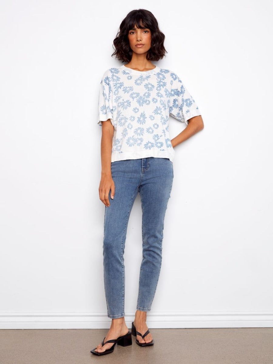 Charlie B Skylar Petal Crochet Short Sleeve Sweater in white with blue floral pattern, paired with blue jeans and black sandals.