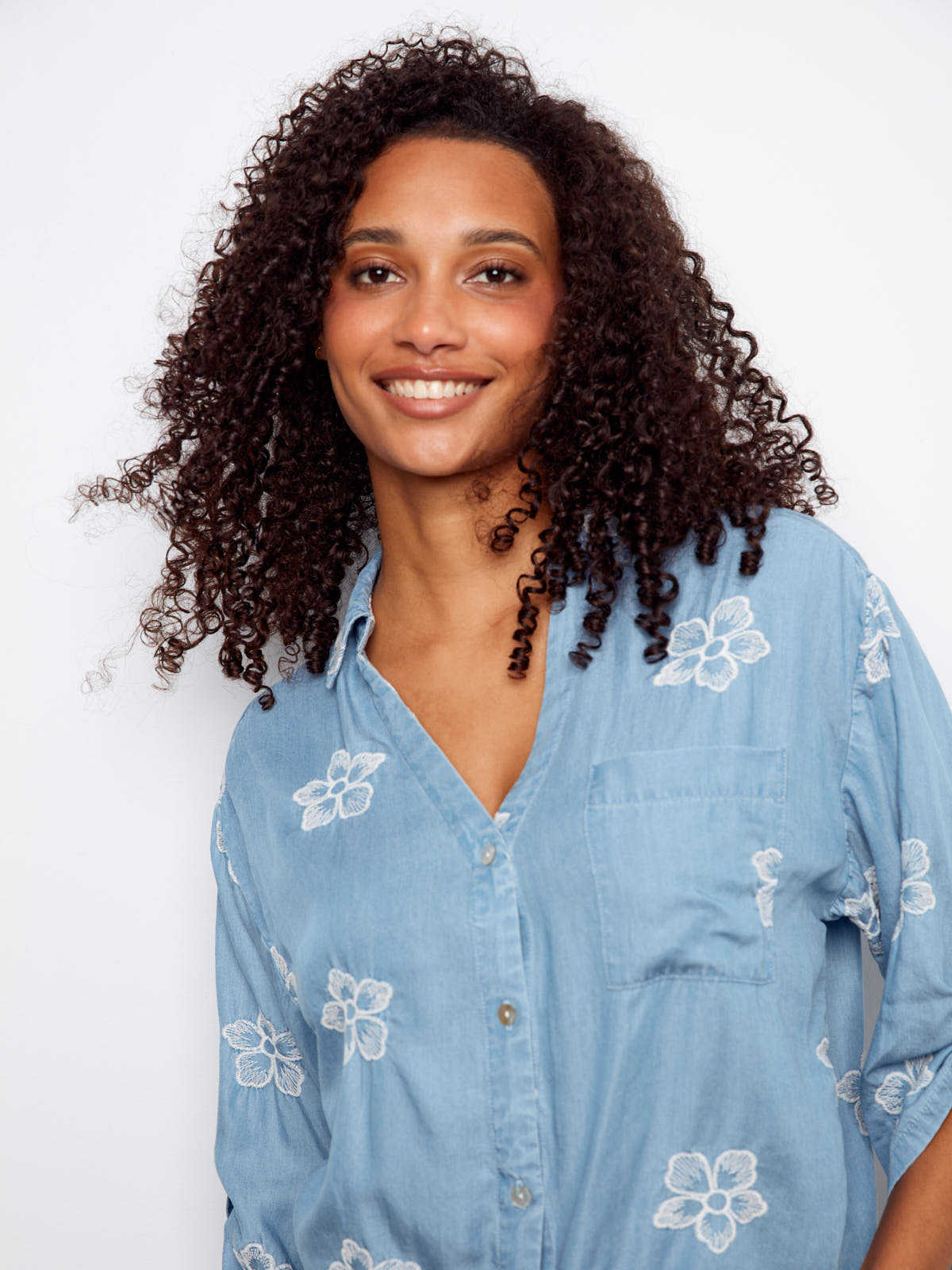 Smiling model wearing a blue Charlie B Embroidered Flower Tencel Button Front Tie Top with white floral embroidery