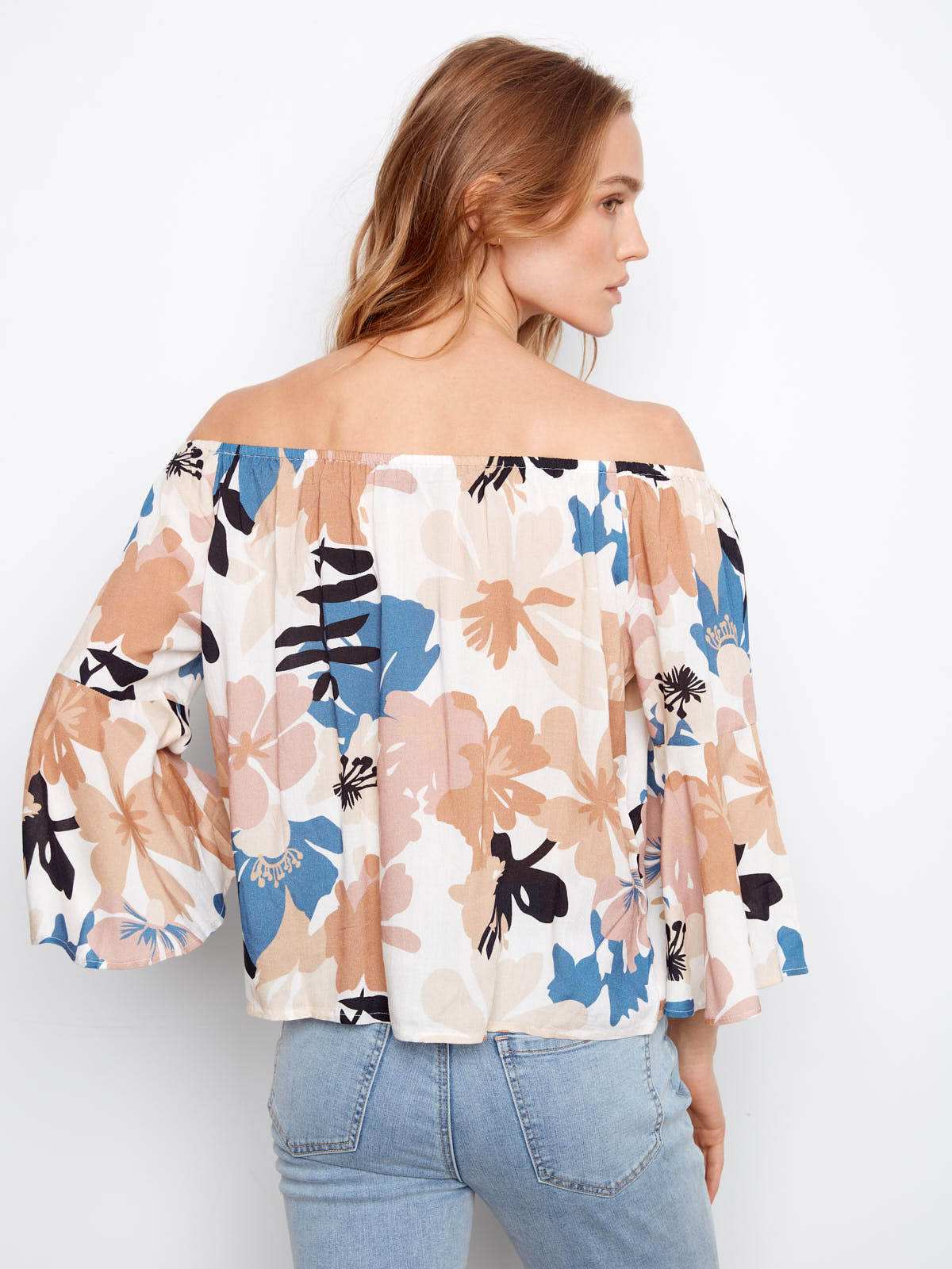 Person wearing an off-shoulder floral top with wide sleeves in neutral and blue tones, paired with light denim jeans.