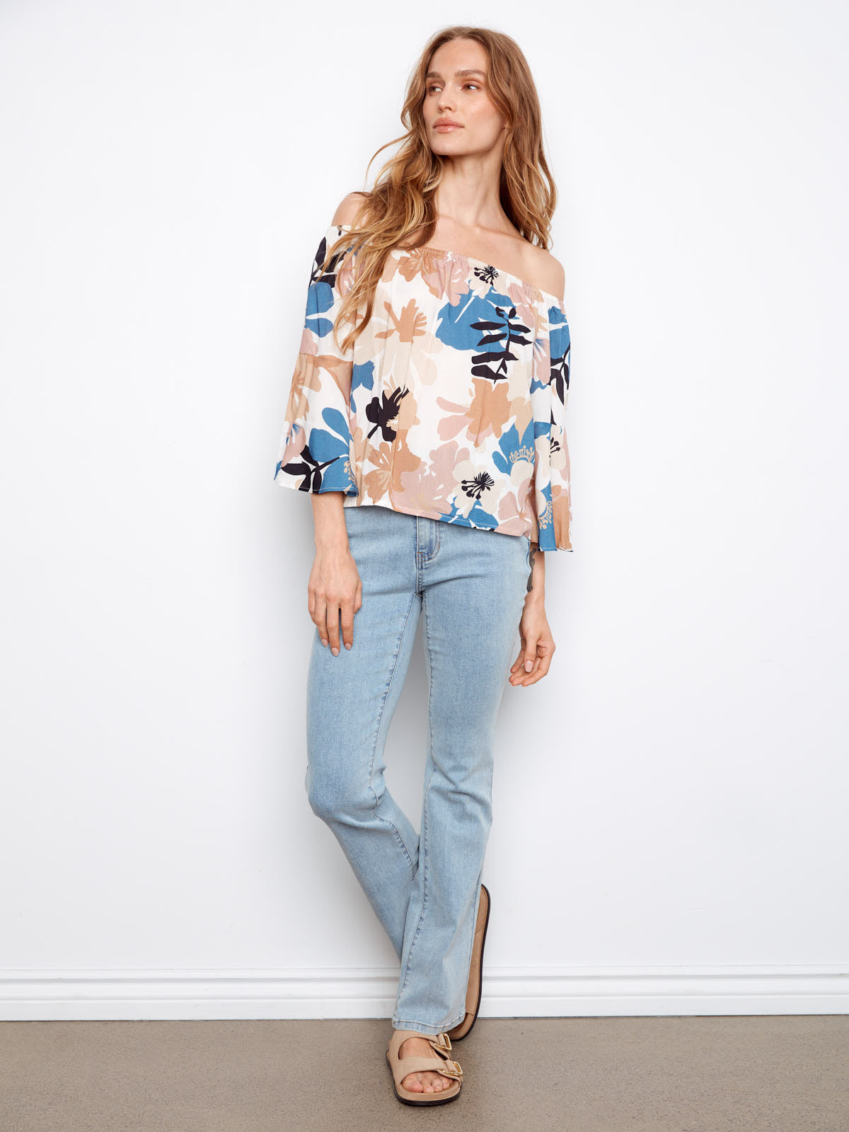 Person in a floral off-shoulder top with blue, beige, and black patterns paired with light blue denim jeans and sandals.