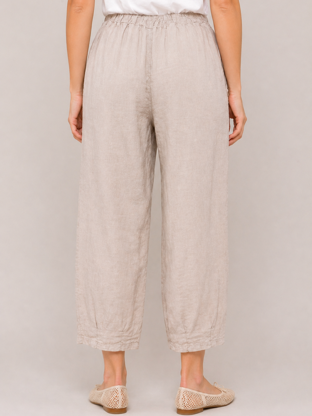Beige loose-fitting linen pants with an elastic waistband and cropped length, paired with light woven slip-on shoes.