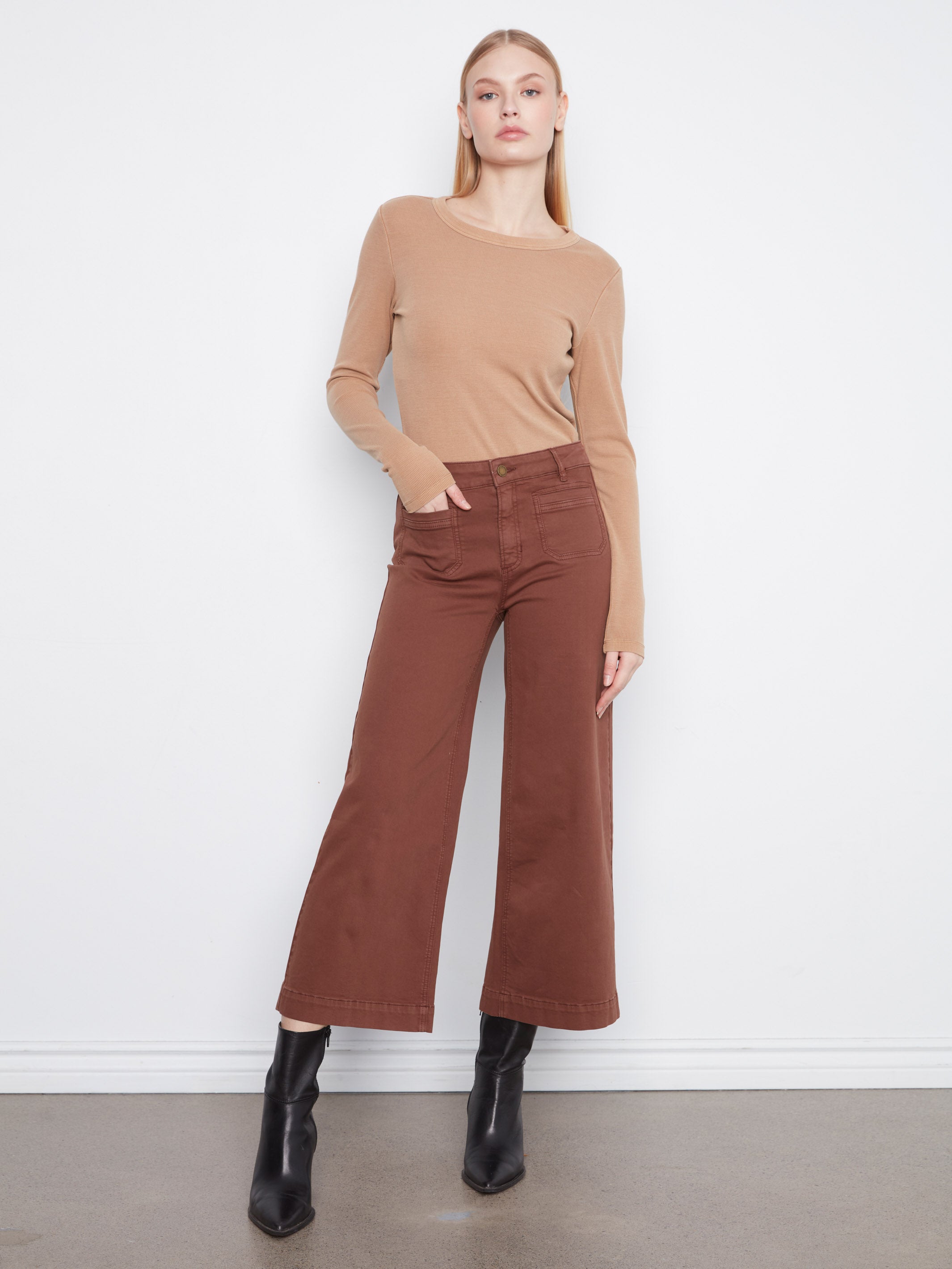 Model in a beige long-sleeve top, wide-leg brown pants, and black ankle boots against a white background.