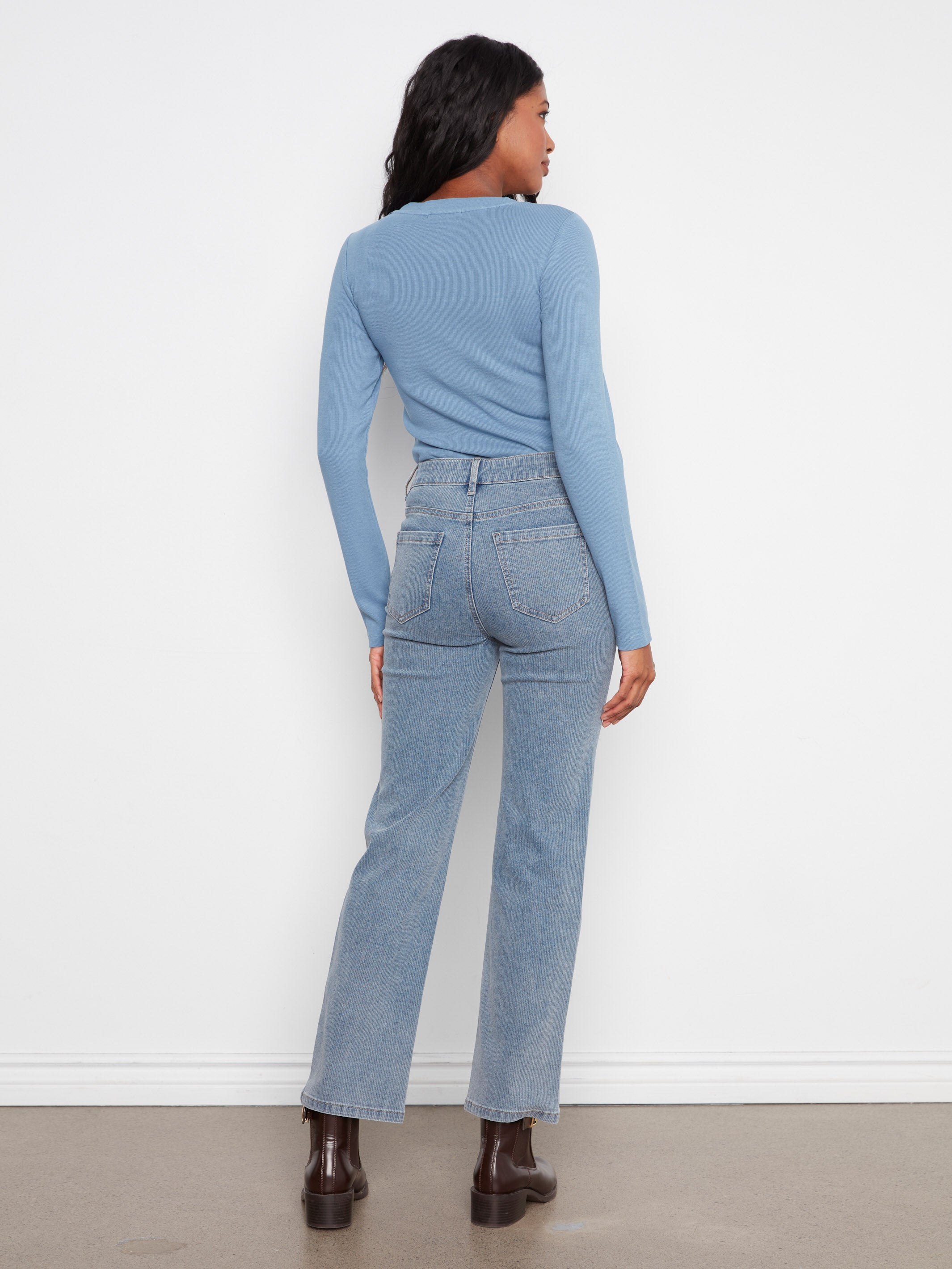 Back view of a figure in light blue long-sleeve top and high-waisted denim jeans standing against a white wall.