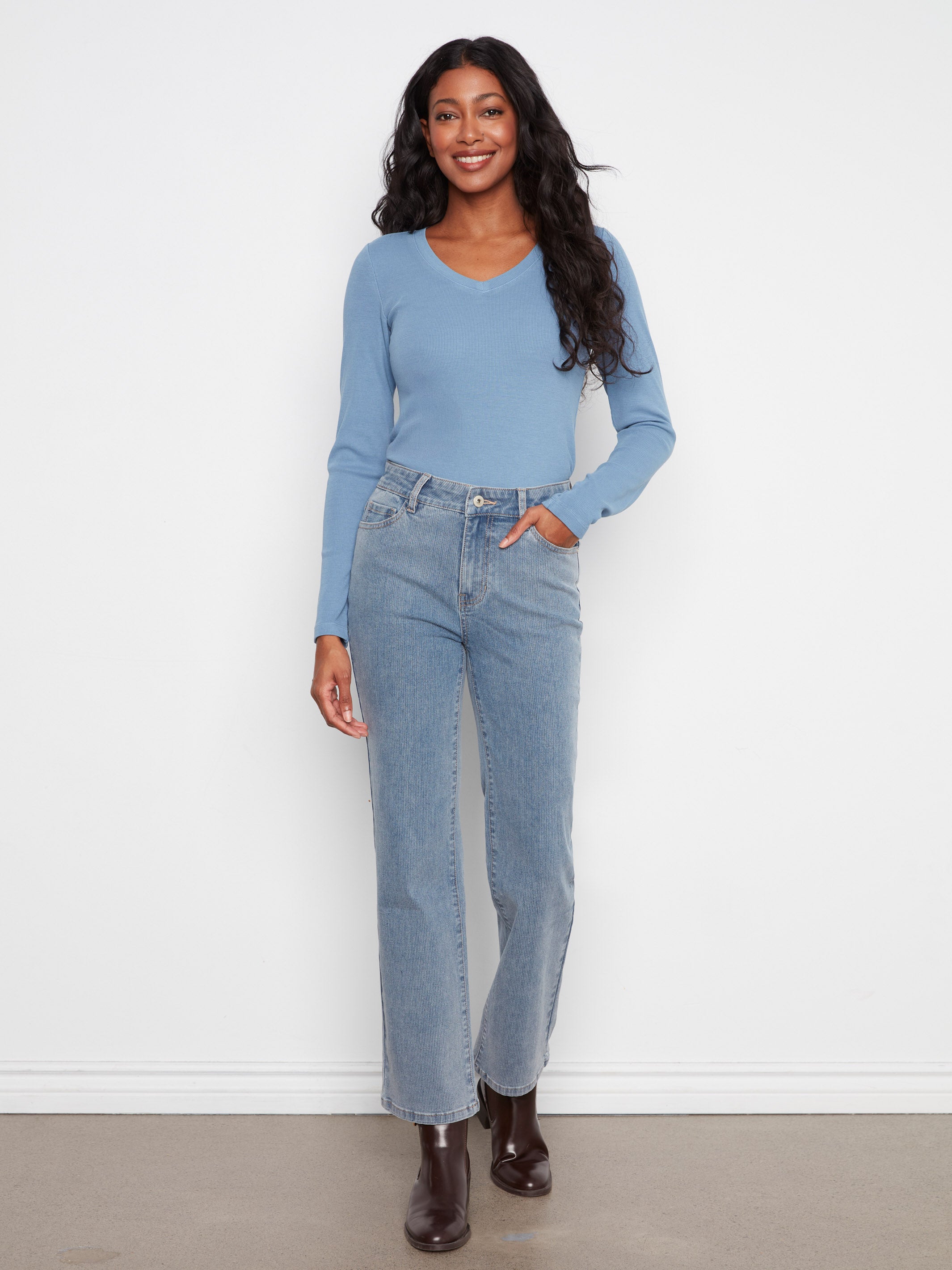 Individual in a light blue long-sleeve top and high-waisted straight-leg blue jeans standing against a white wall.