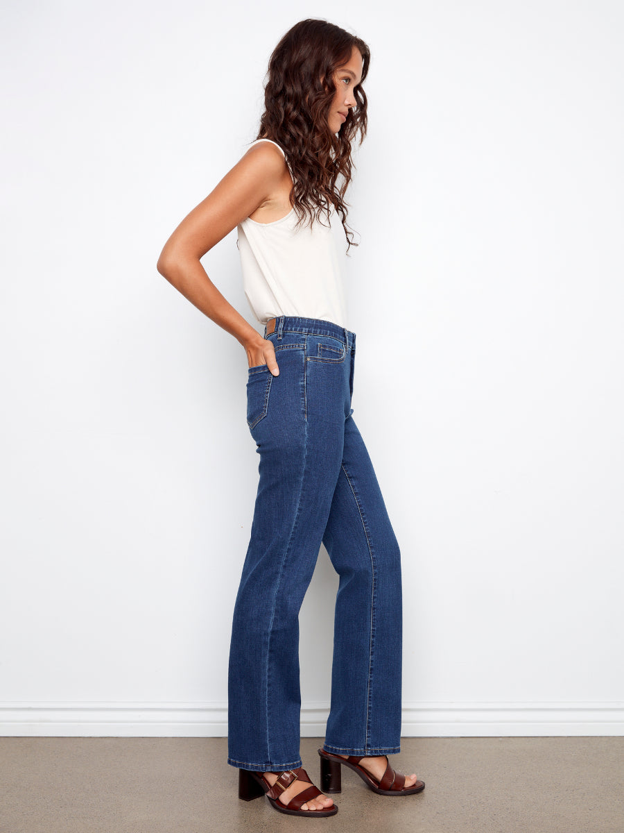 Side view of a model wearing Charlie B Charlotte Straight Leg Denim Jeans in indigo with a white sleeveless top and brown sandals