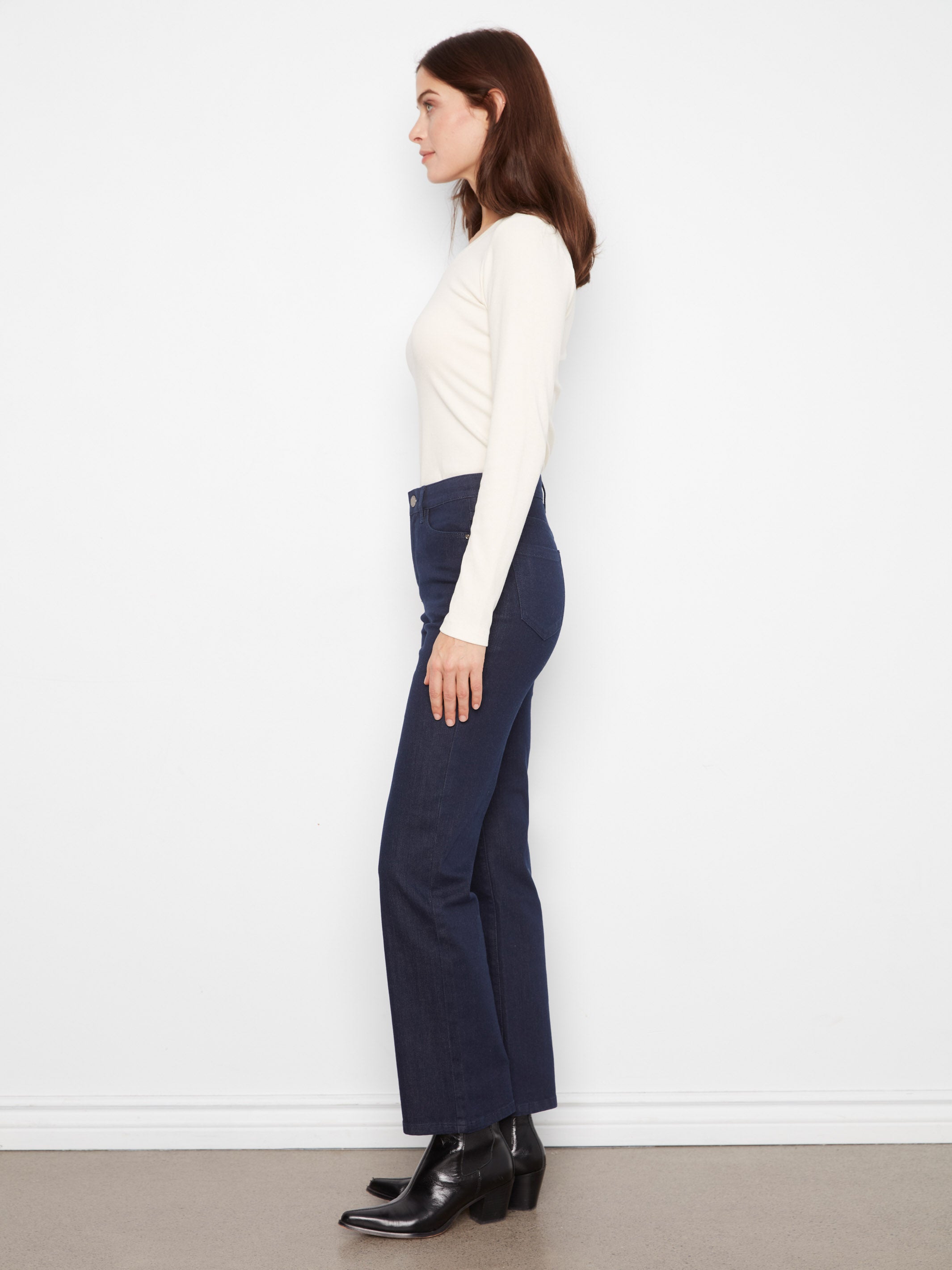 Side view of a figure in a cream long-sleeve top, dark blue denim jeans, and black ankle boots against a white wall.