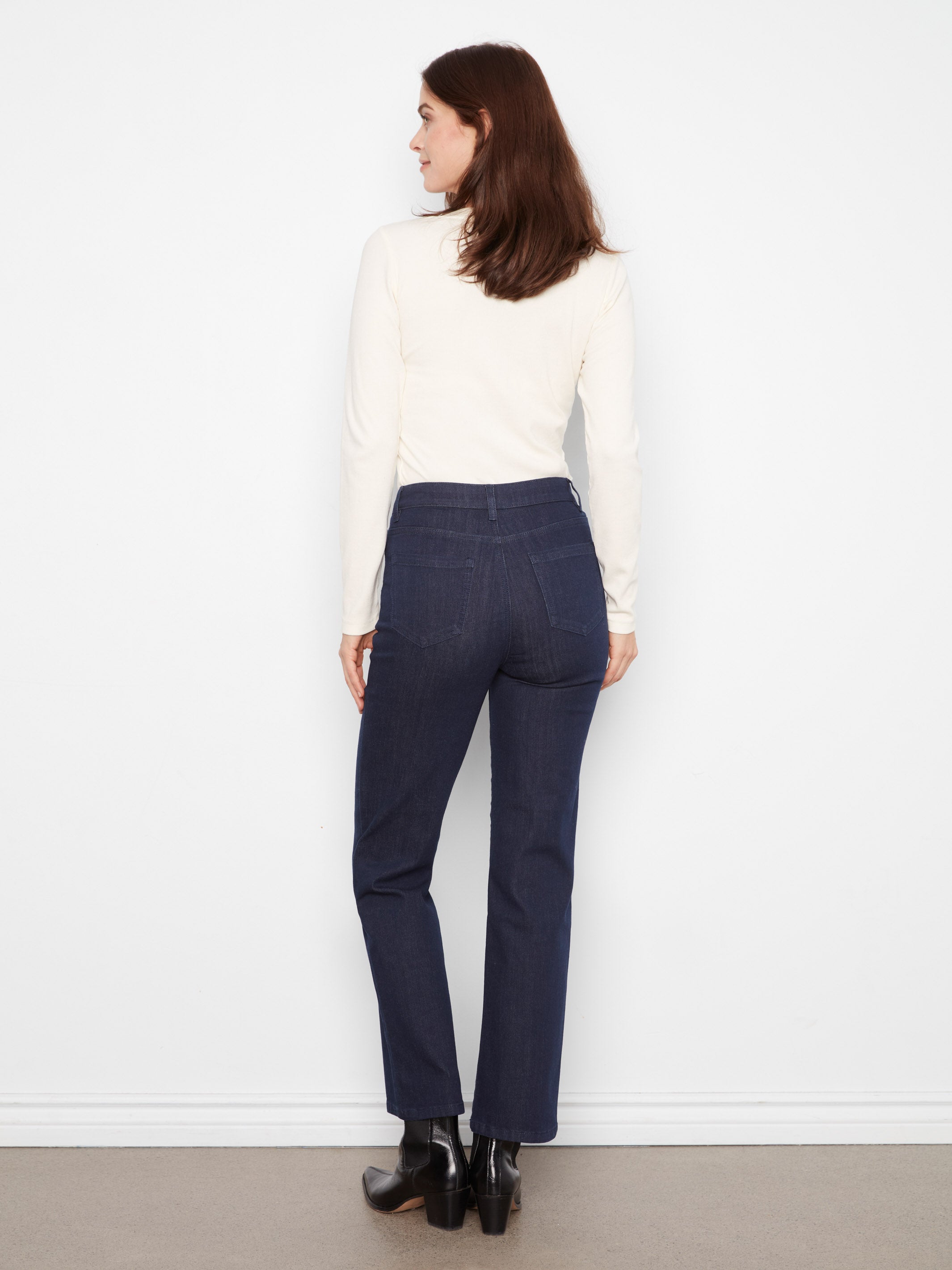 Back view of denim jeans paired with a long-sleeve cream top and black ankle boots against a white background.