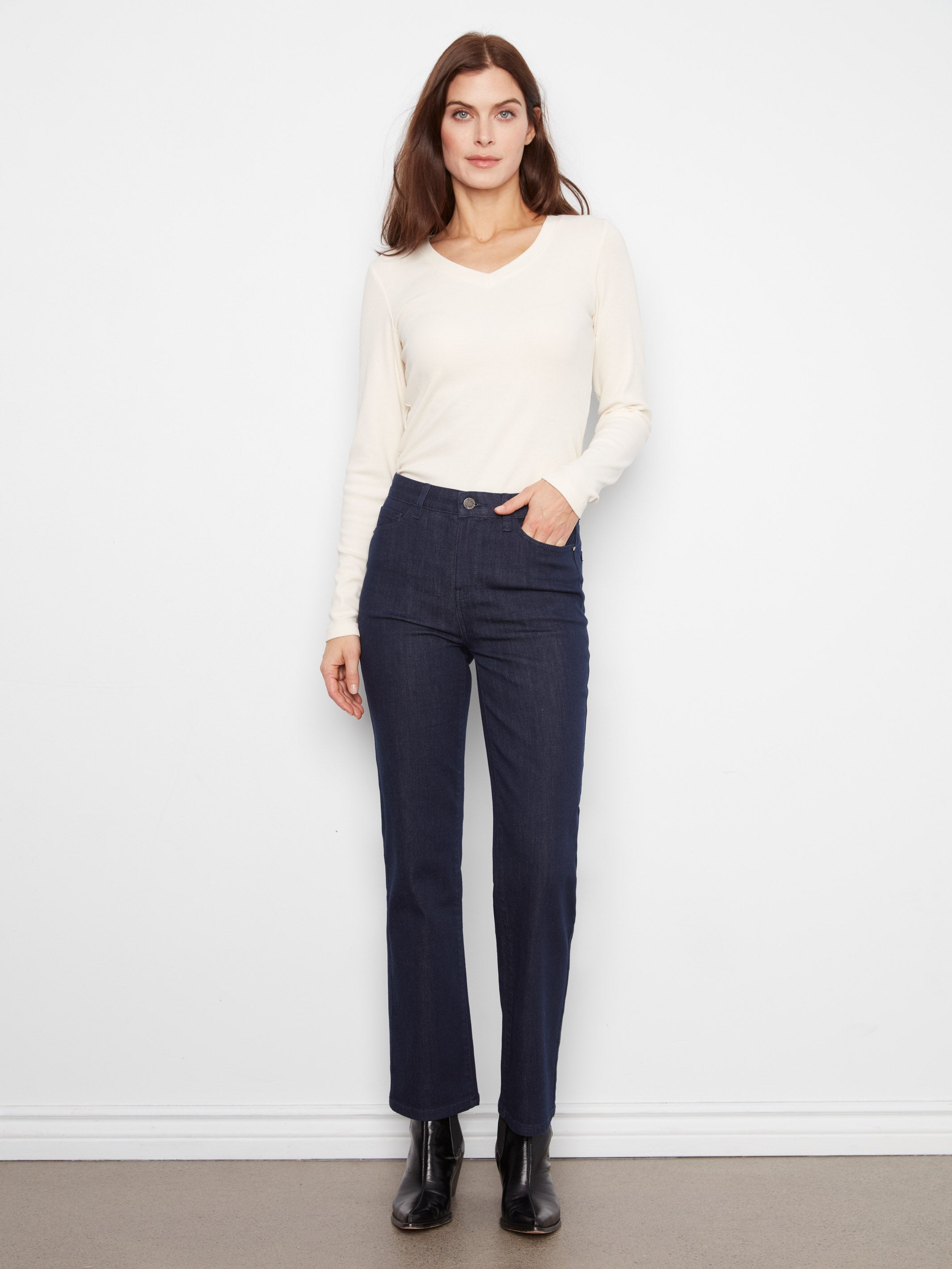 Slim fit dark denim pants paired with a cream long-sleeve V-neck top and black ankle boots against a white background