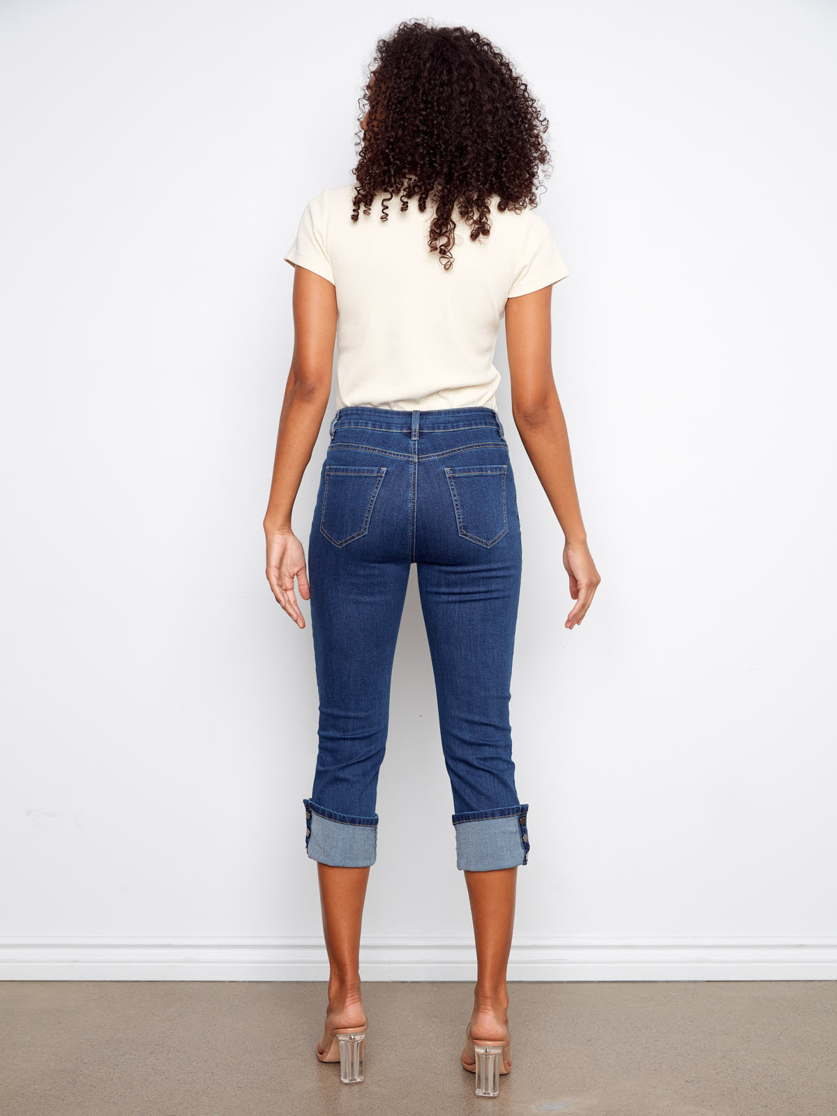 Back view of Charlie B Up Down Snap 26” Cropped Denim Capris in indigo blue worn with a neutral top and clear heels