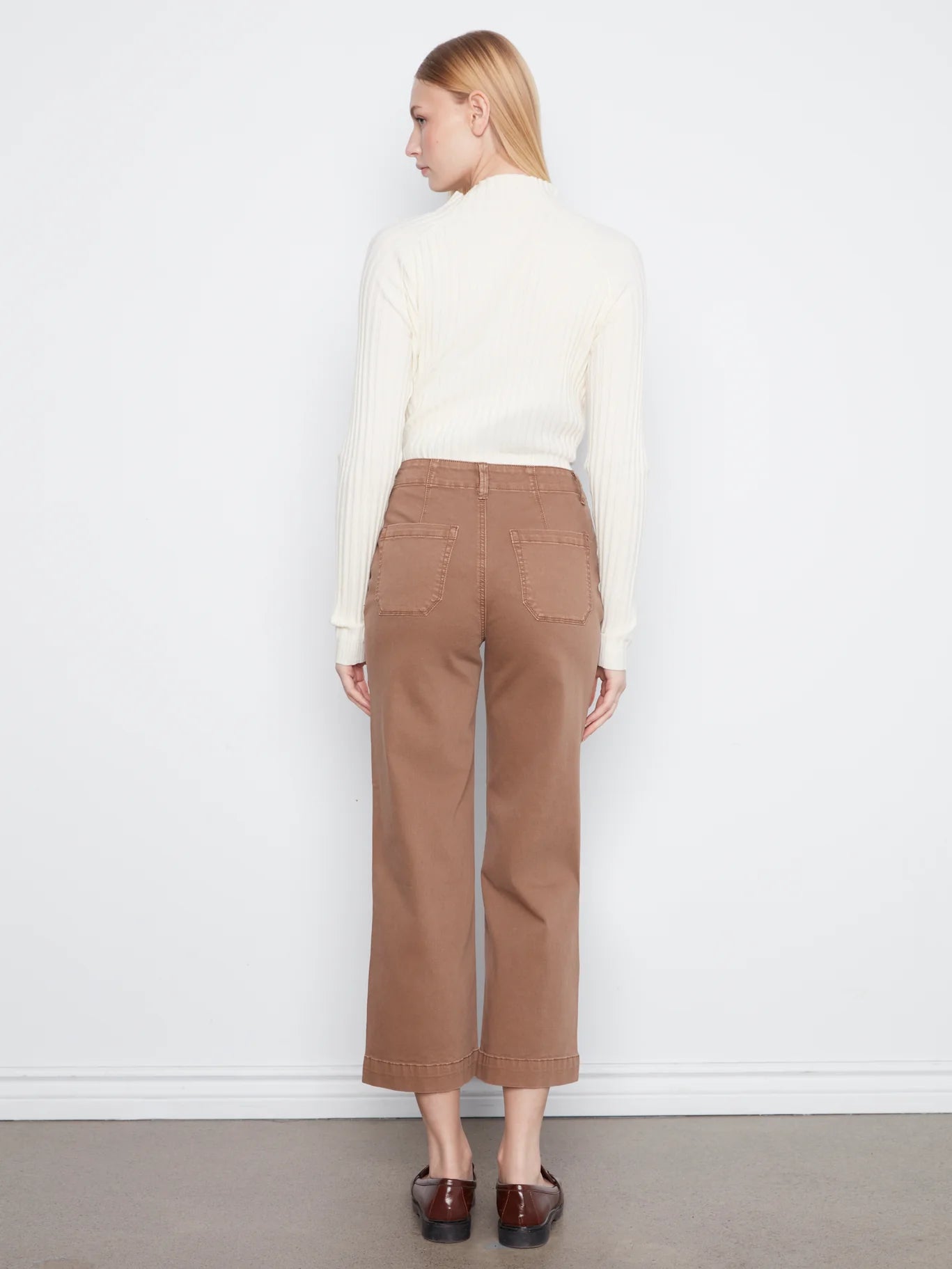 Back view of a figure in a white ribbed sweater and cropped brown denim pants against a plain white background.