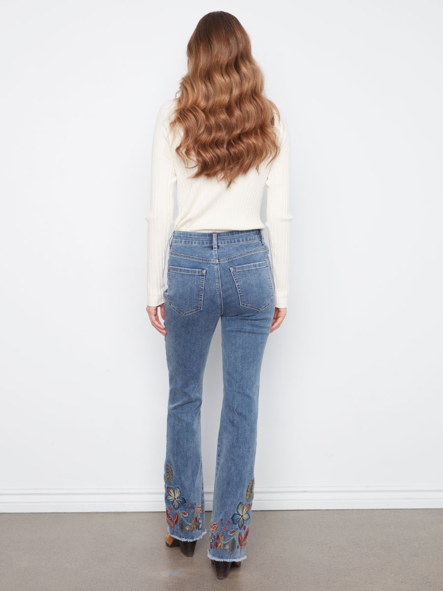 Back view of medium blue denim jeans with floral embroidery on the lower legs paired with a white ribbed long sleeve top.