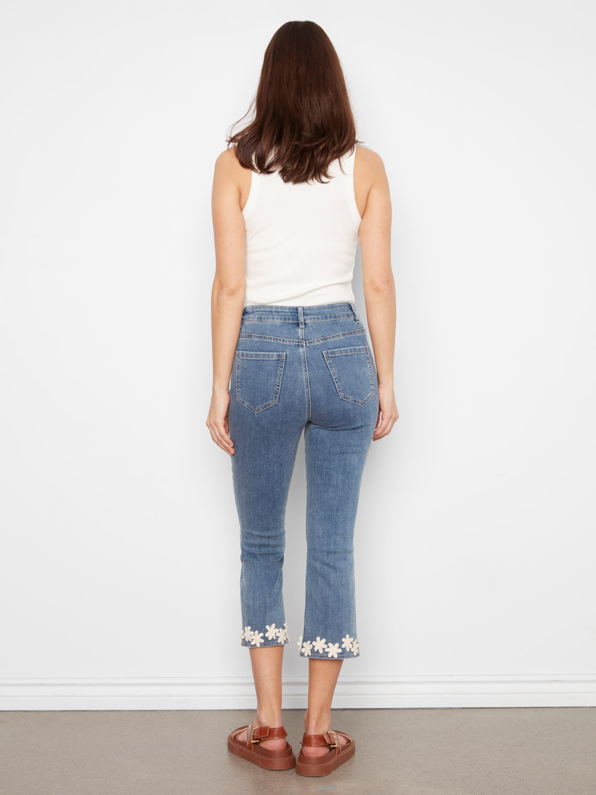 Back view of Charlie B Crochet Flower Hem Denim Capris in medium blue denim with floral crochet trim and brown sandals.